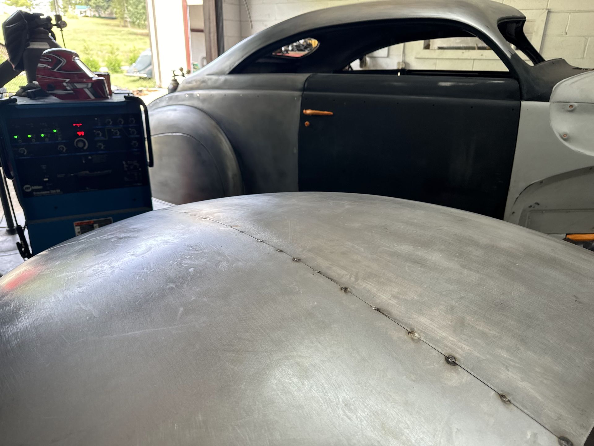 Inside a vintage car body, showing a gray primered roof and dashboard near a workshop setup