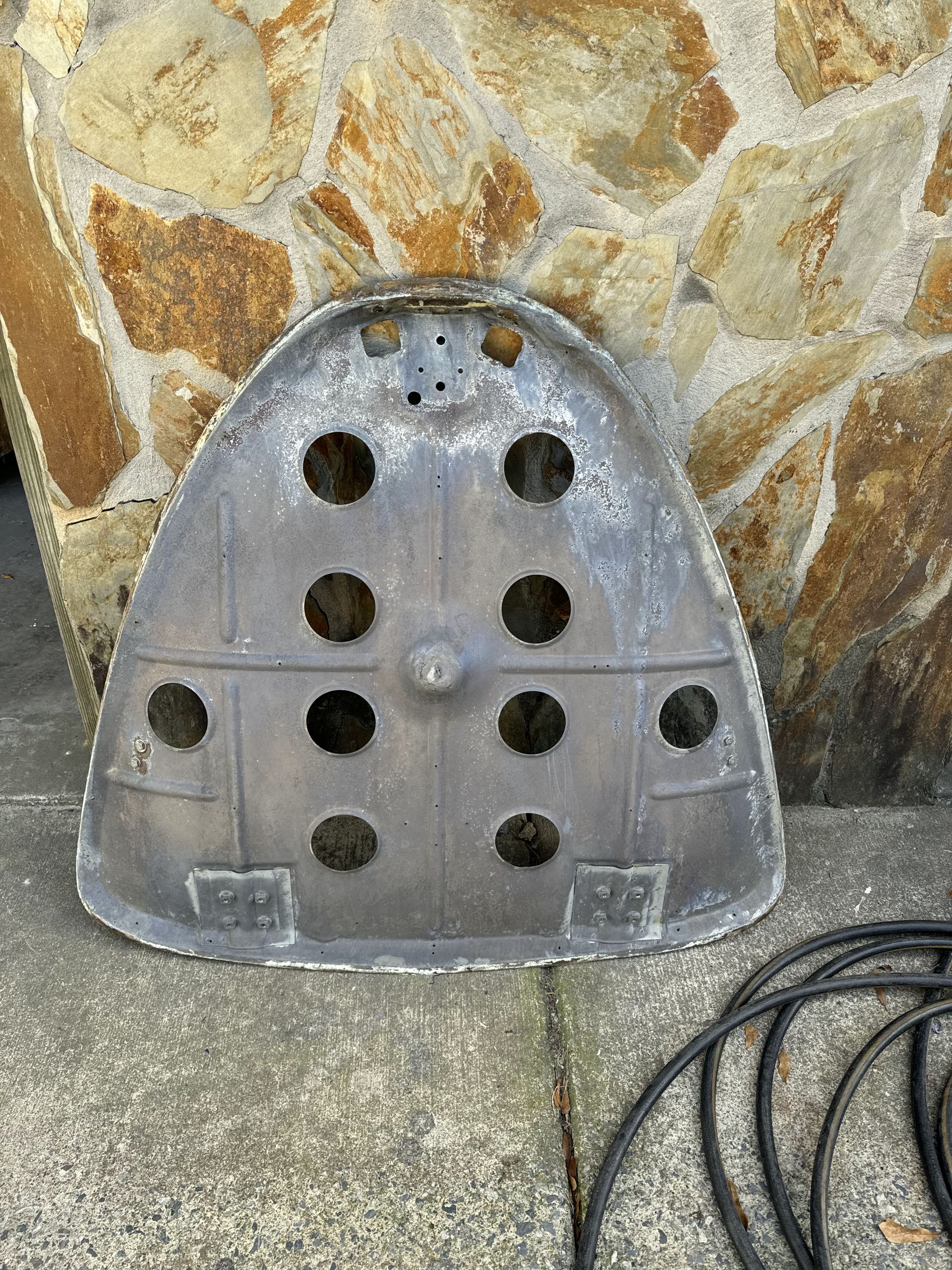 Gray plastic panel with multiple round holes leaning against a stone wall on concrete, with a black cable nearby