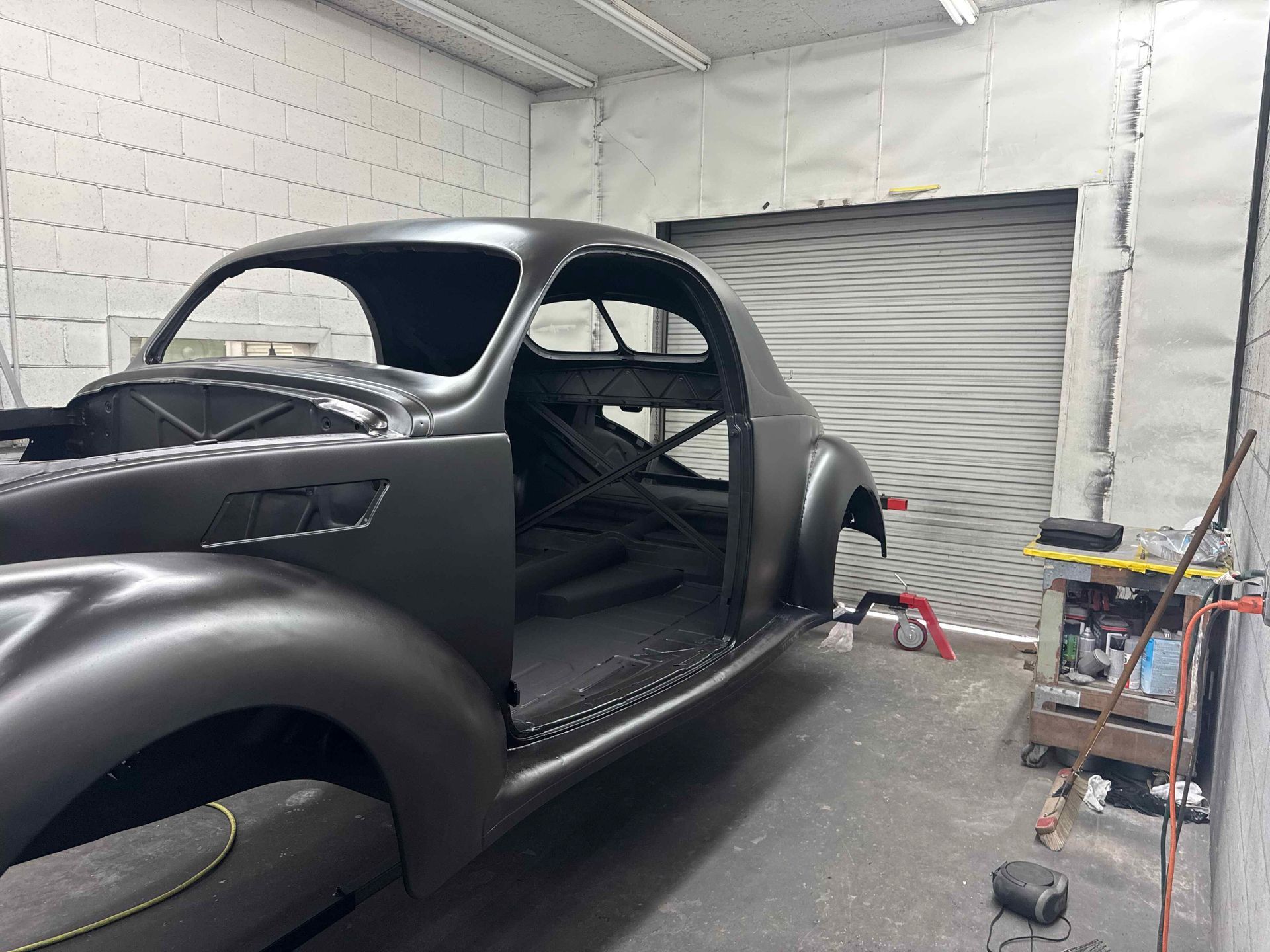 Matte black classic car body in a workshop, missing windows and wheels, with tools nearby