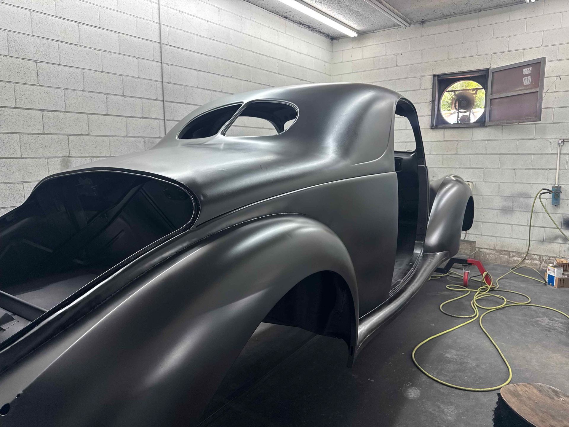Matte black classic car body in a garage, with masked windows and sanding tools nearby.