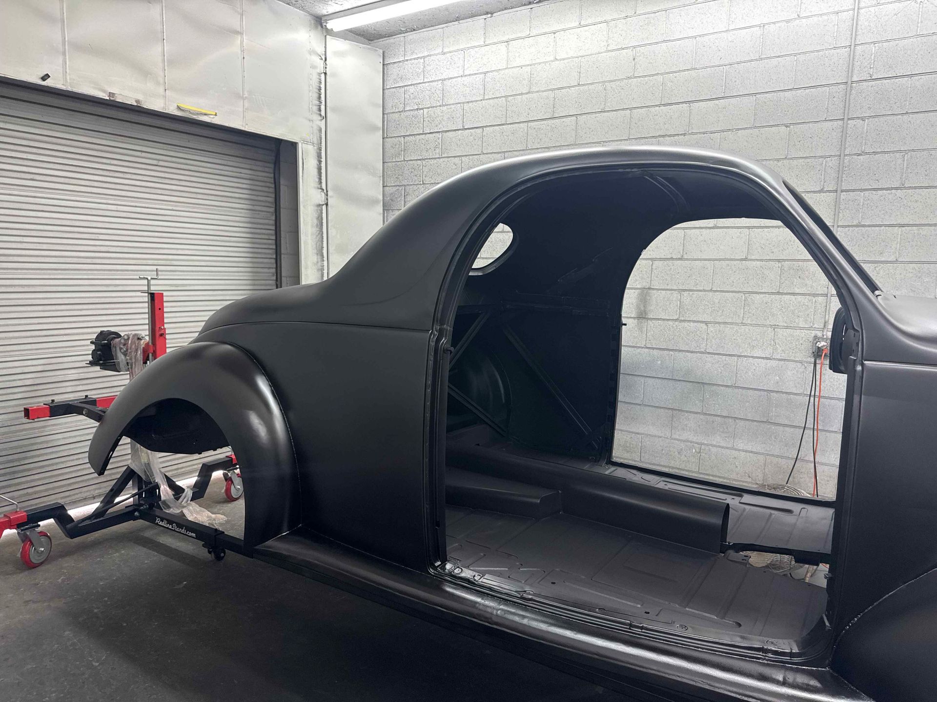 Black car body shell in a garage, mounted on a chassis with a roll cage visible.