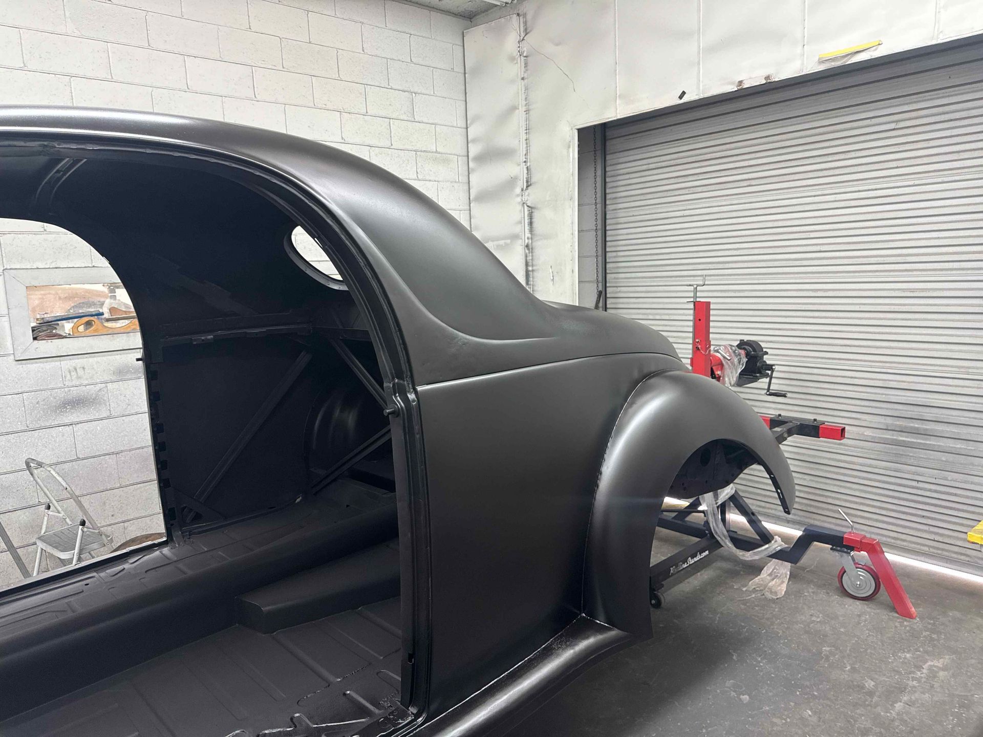Black classic car body shell in a workshop, with rear fender and wheel assembly visible.