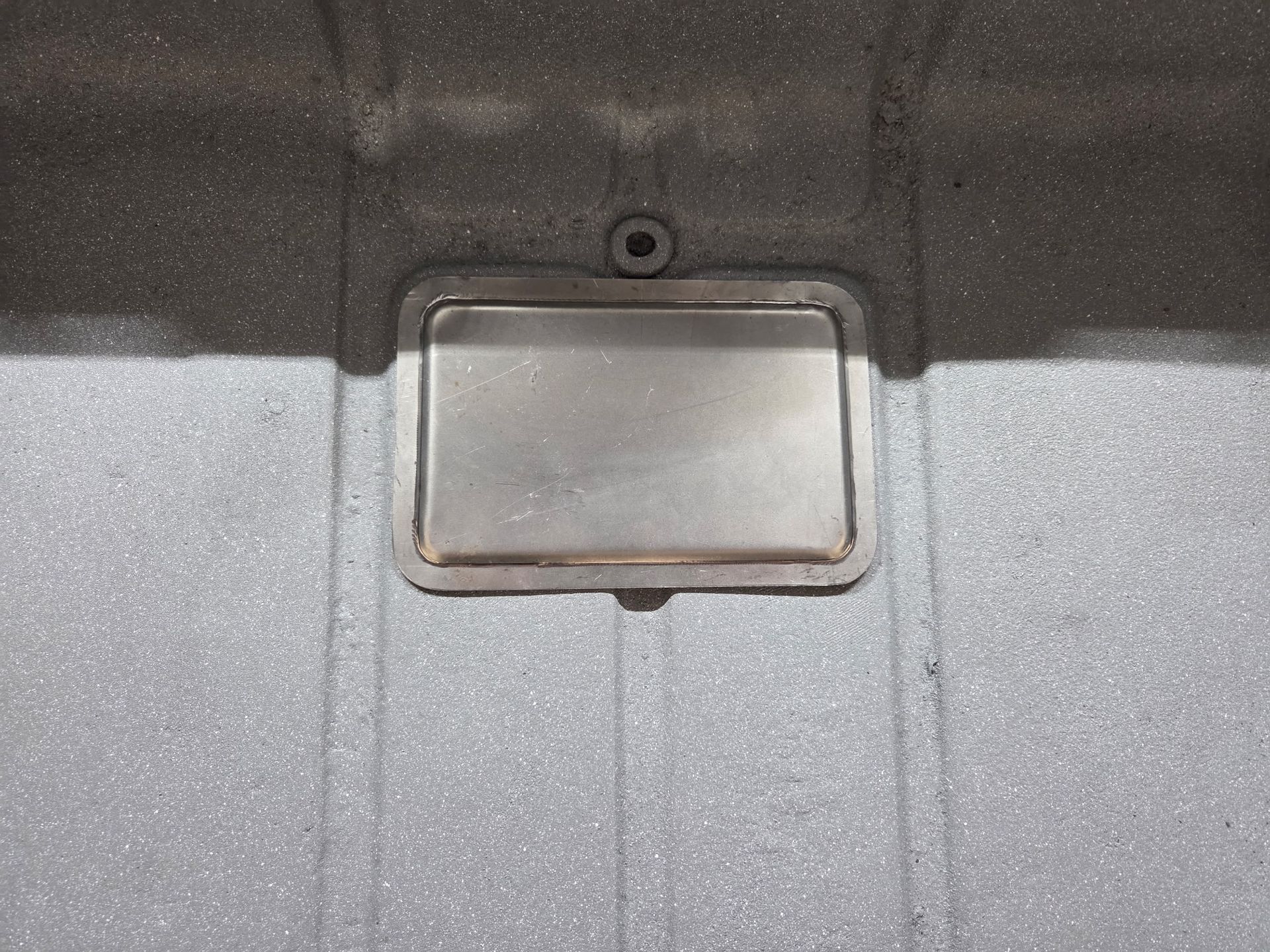 Rectangular metal access hatch on a gray textured surface with a small circular opening above it