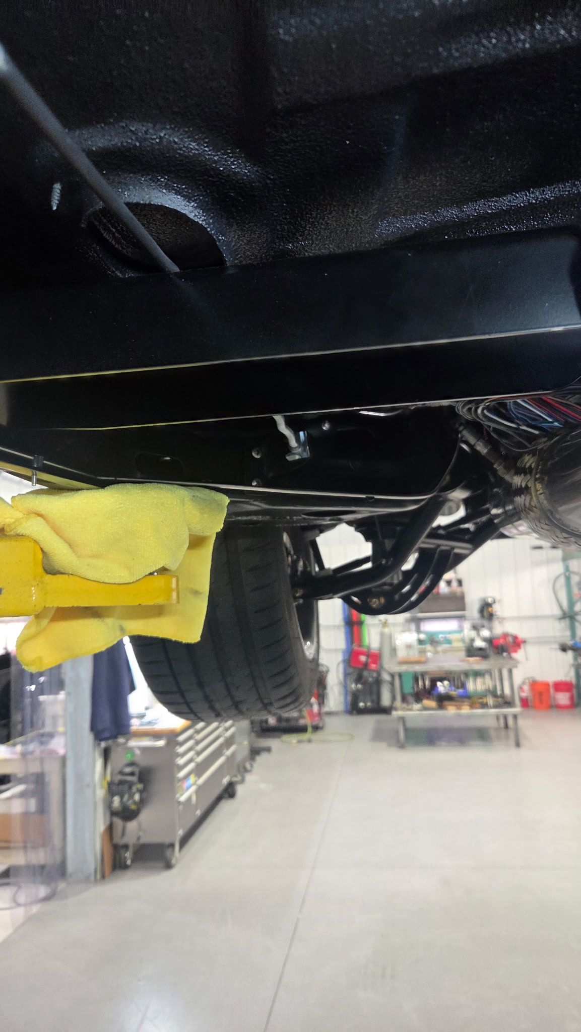 Undercarriage of a black vehicle on a lift in an auto repair shop, with a yellow lift arm visible