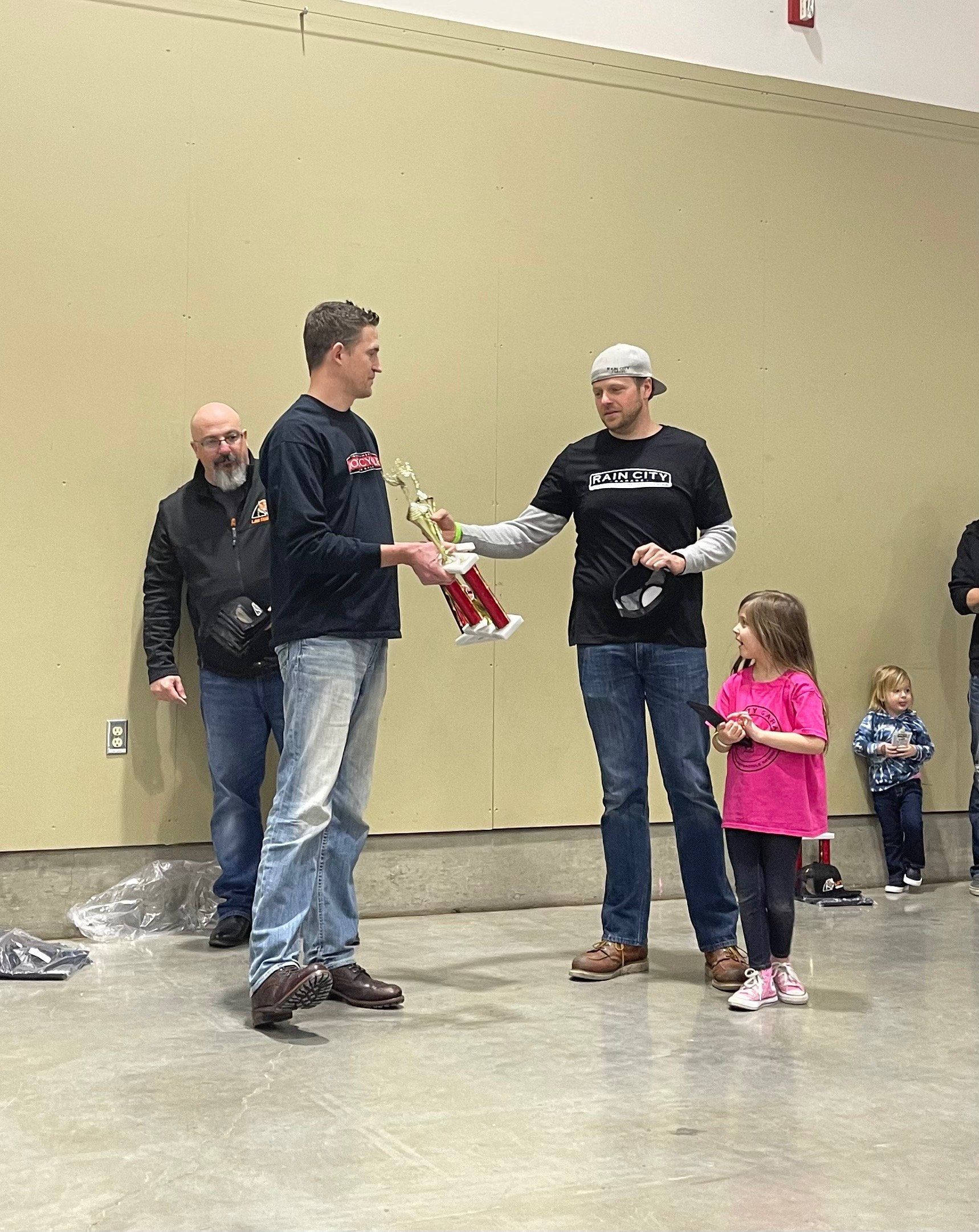 Three people and two children stand in a hall, one man handing over a red item while others watch.