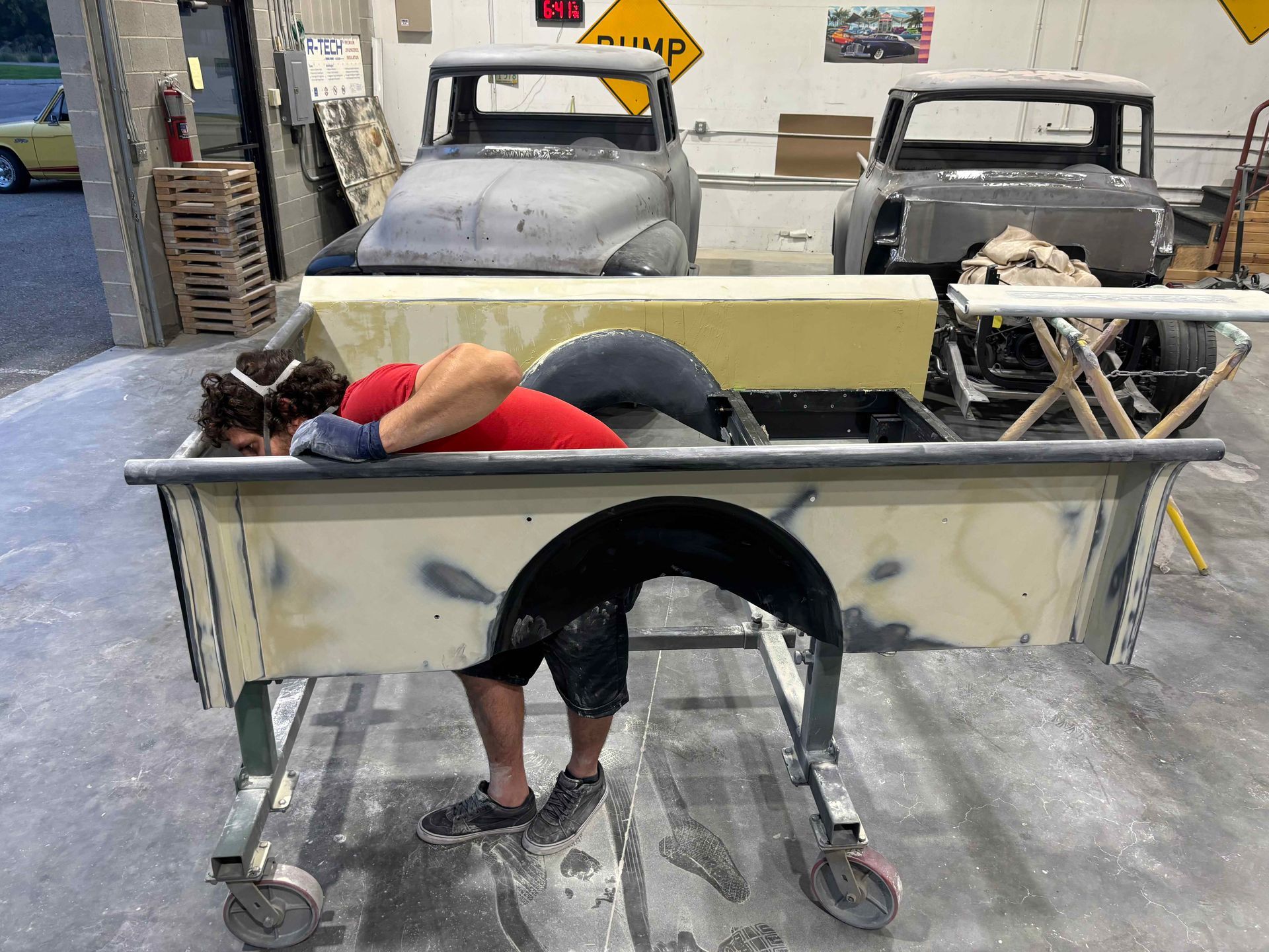 Person bent over a large metal cart in a workshop, with two old cars in the background.