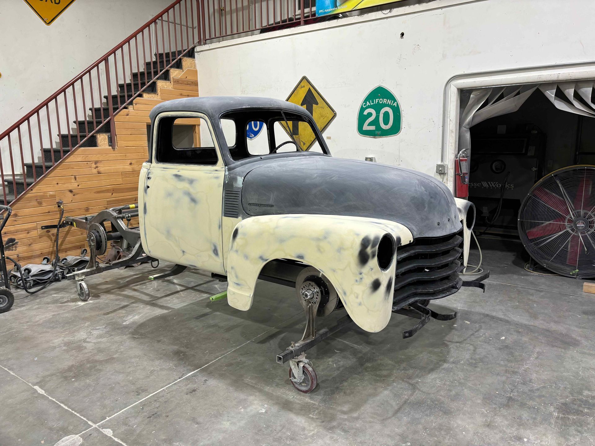Partially restored vintage truck cab with cream primer in a garage, front on stands beside stairs and tools