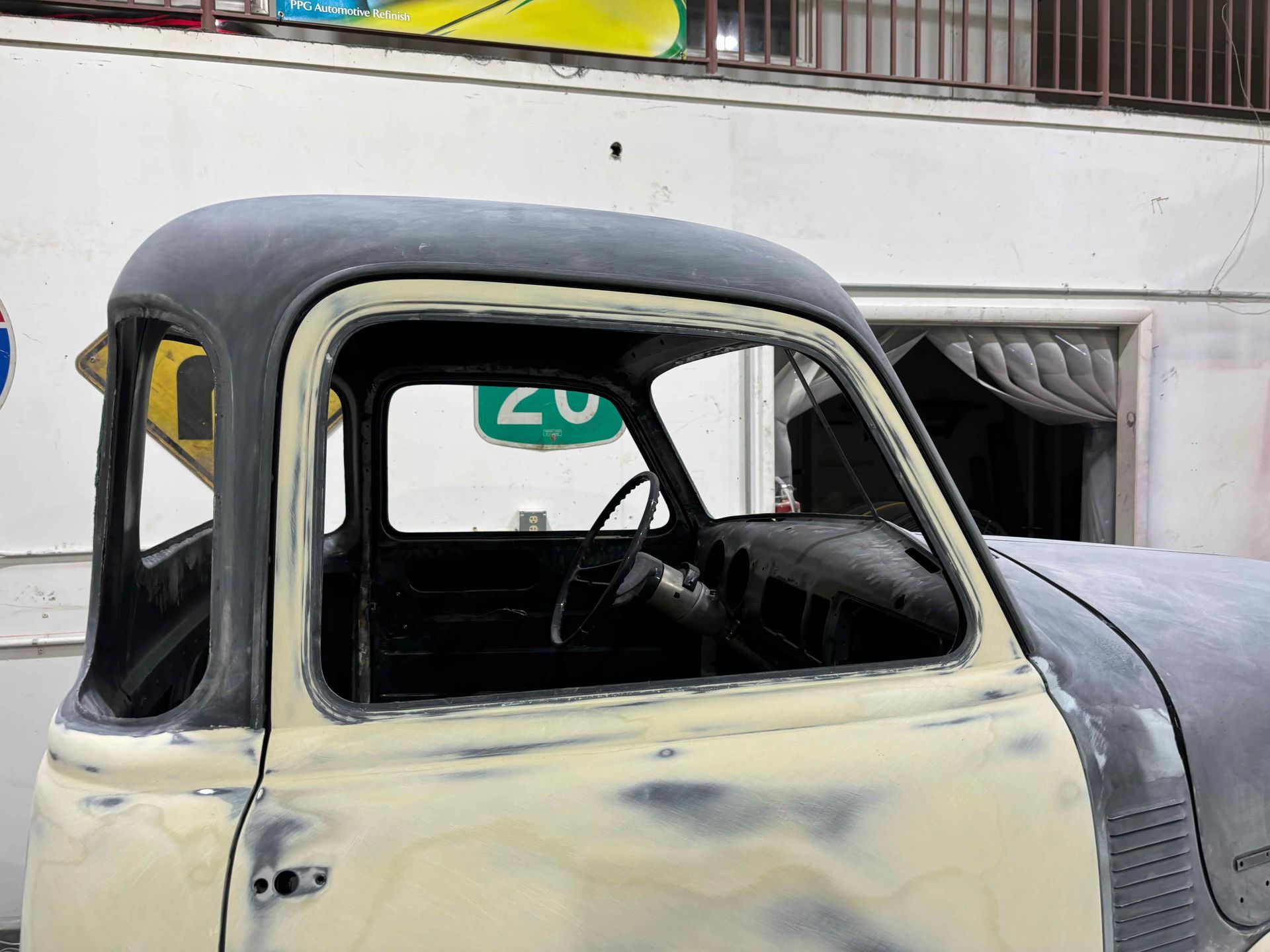 Old cream-colored vintage truck cab with a black roof tarp parked in front of a building.