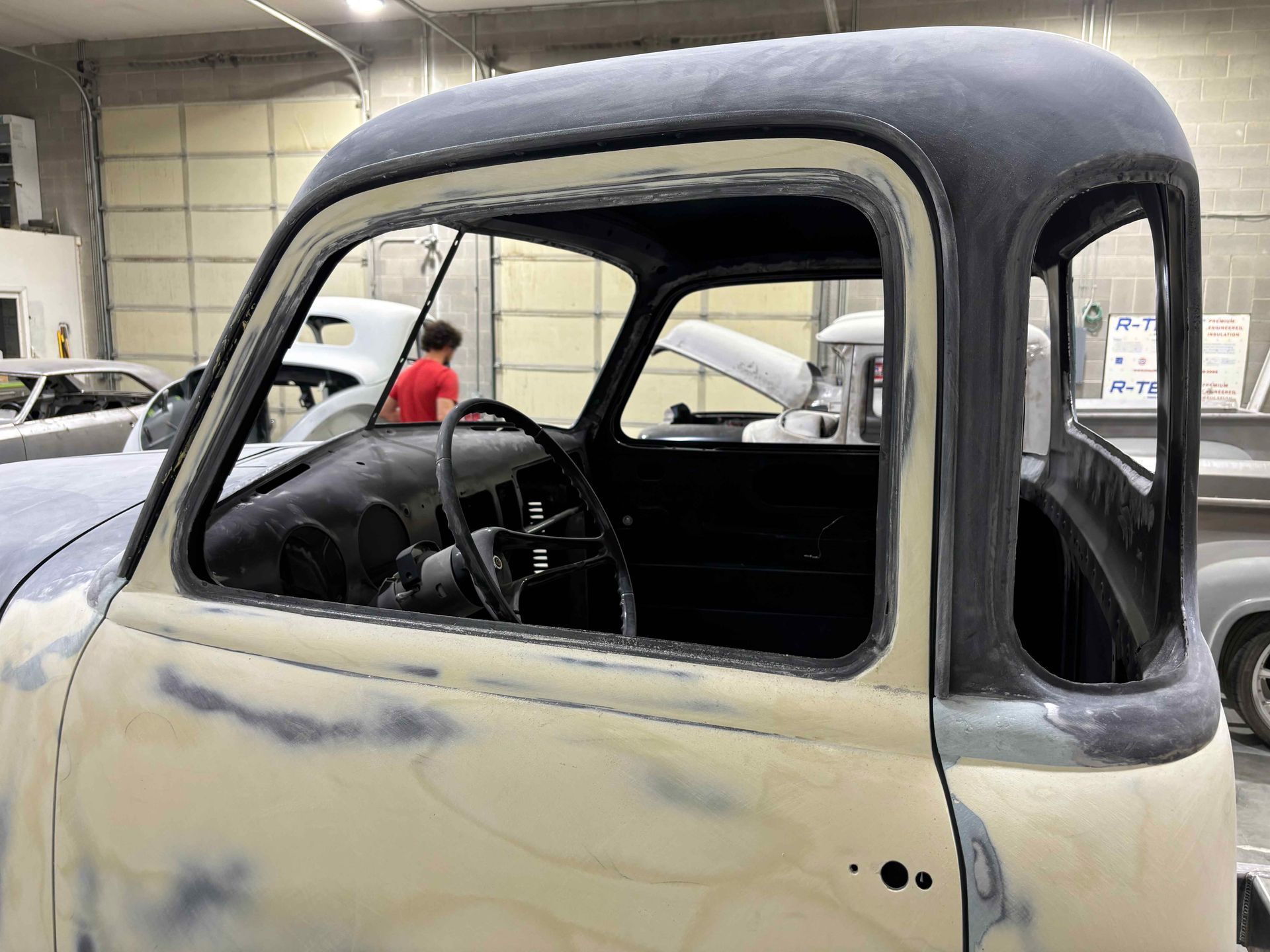 Partially restored vintage truck cab in a garage, with primer-gray body and black roof.