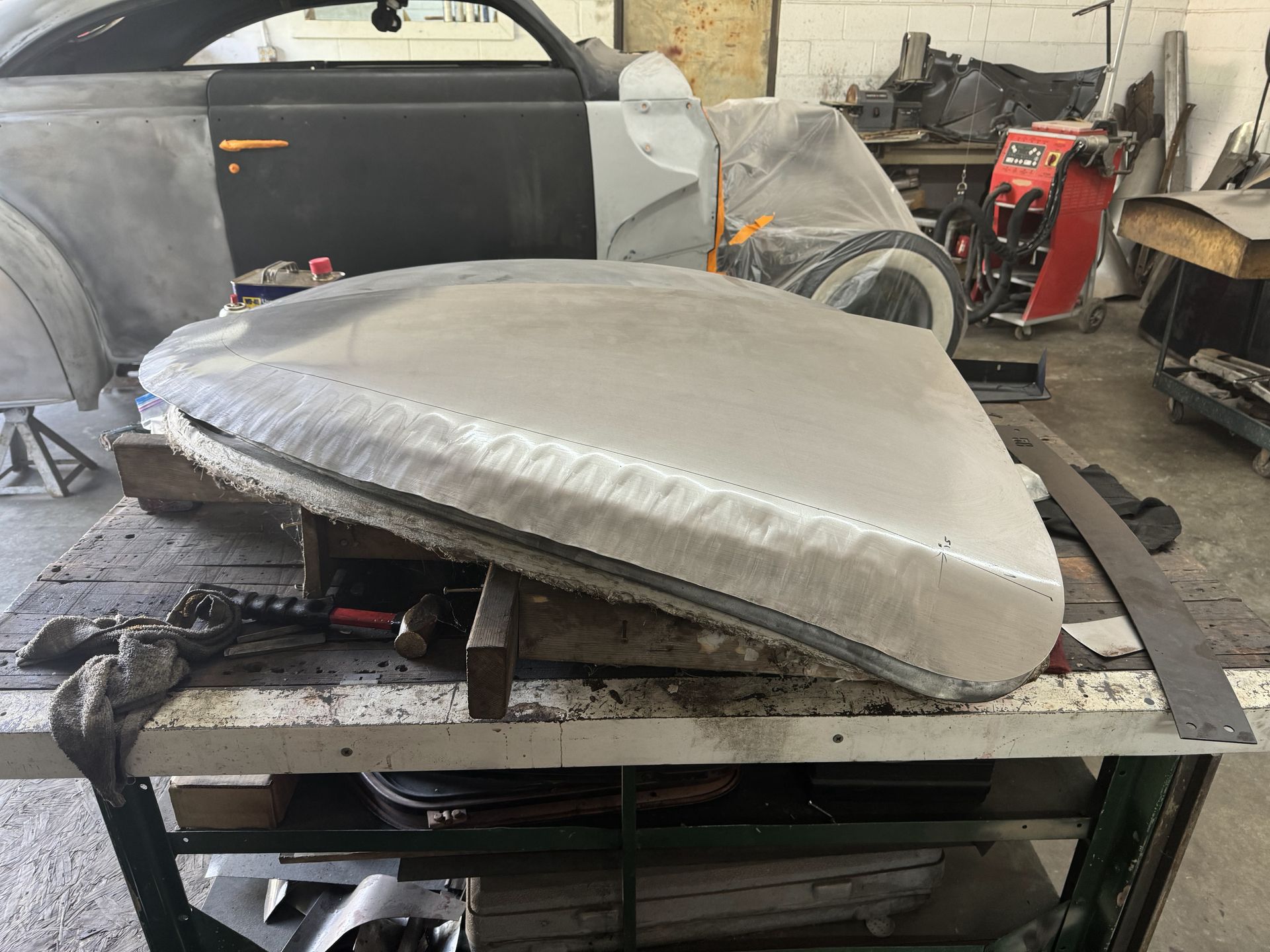 Primed car body panel on a metal worktable in an auto shop, with a classic car shell in the background