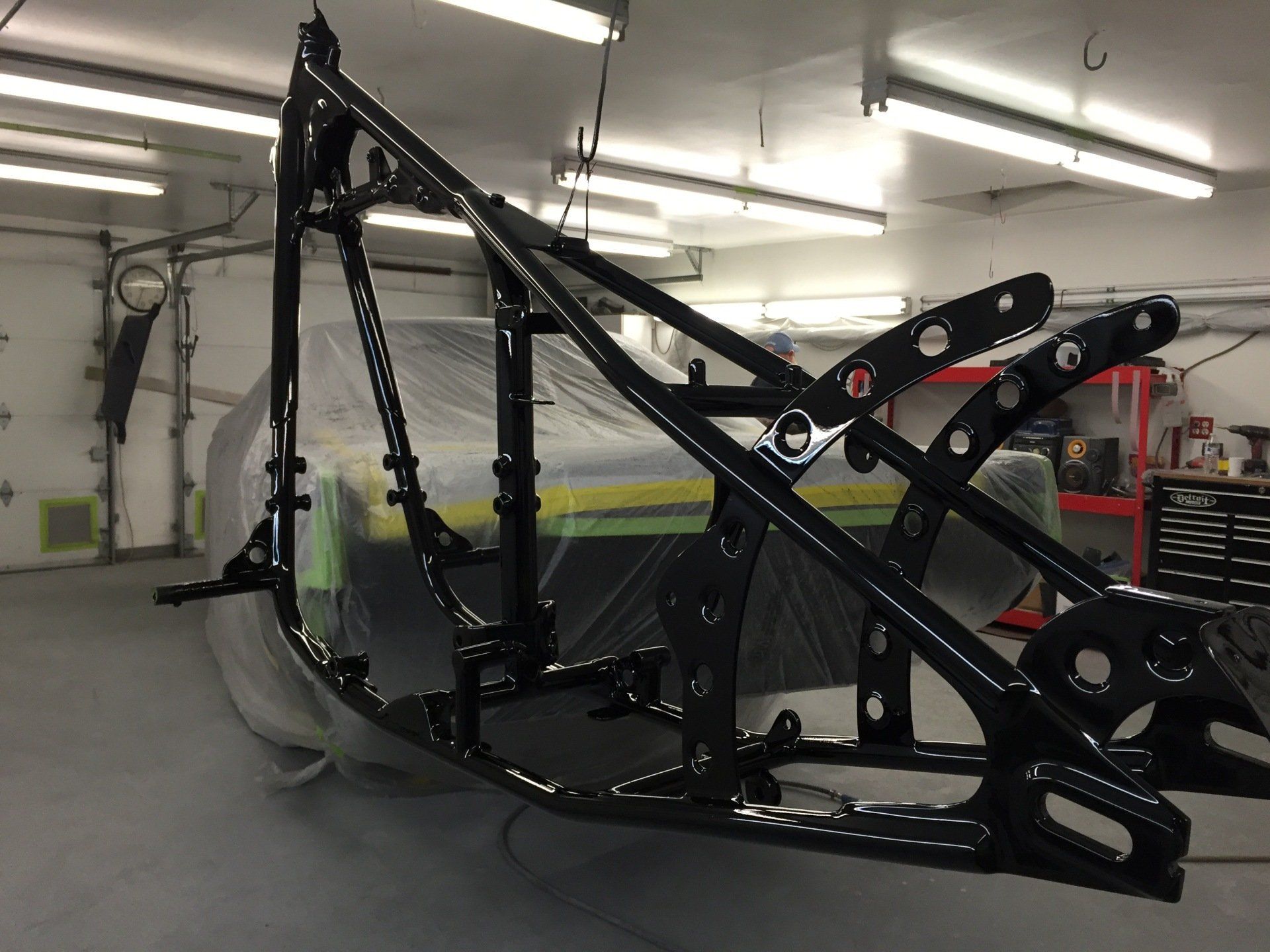 Black metal excavator bucket frame in a workshop, shown from the side under fluorescent lights.