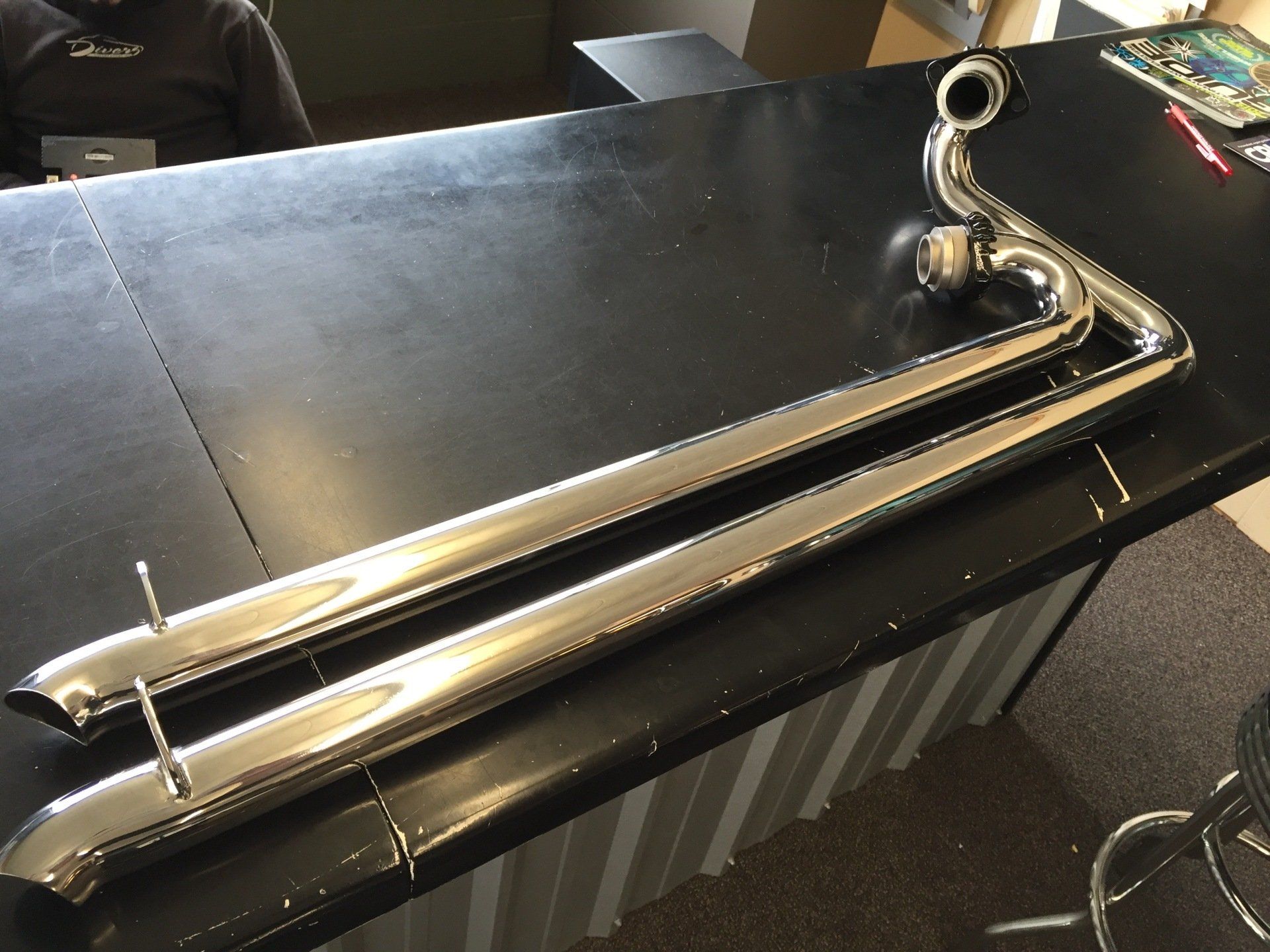 Chrome motorcycle exhaust pipes on a black table, with curved mounting ends visible.