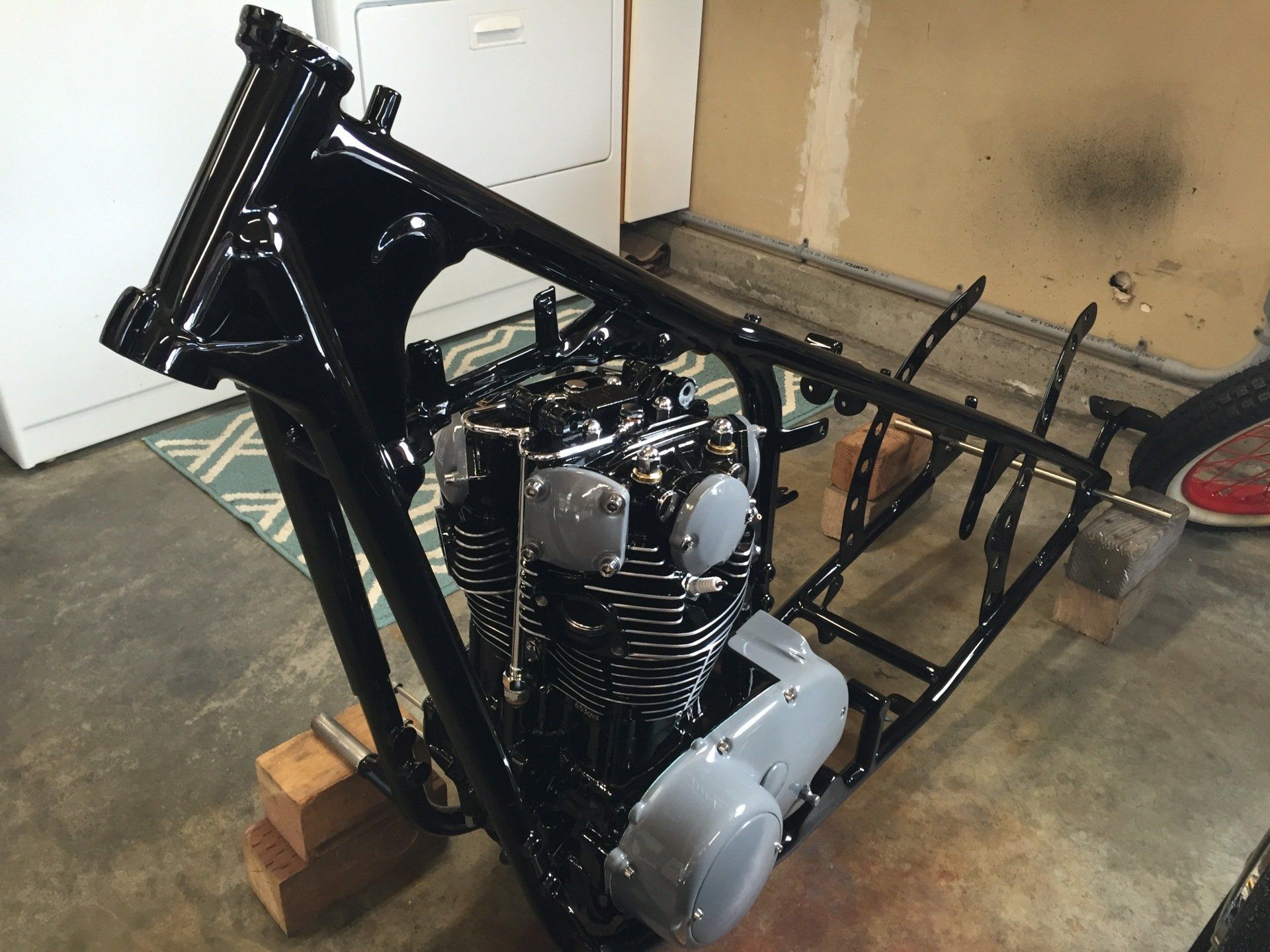Black motorcycle frame with engine in a garage, resting on bricks, beside a detached rear rack