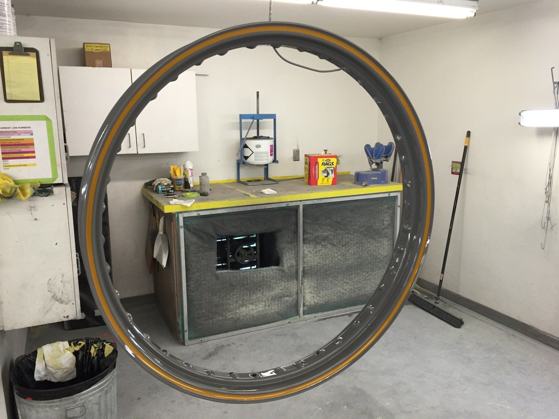 Large bicycle wheel standing in a workshop garage, with tools and supplies on a workbench behind it.