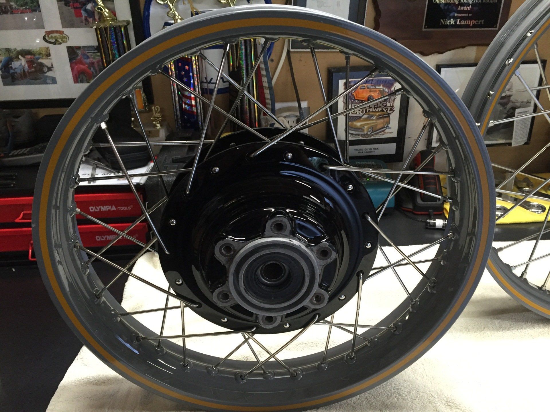 Black motorcycle wheel with spokes and tire, shown close-up in a garage or workshop.