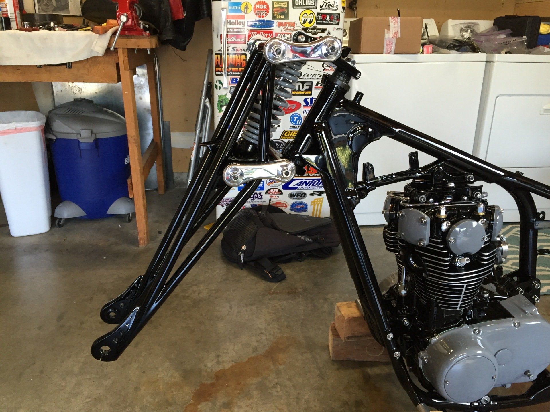 Black motorcycle frame and engine in a garage, with chrome front forks and scattered workshop items nearby