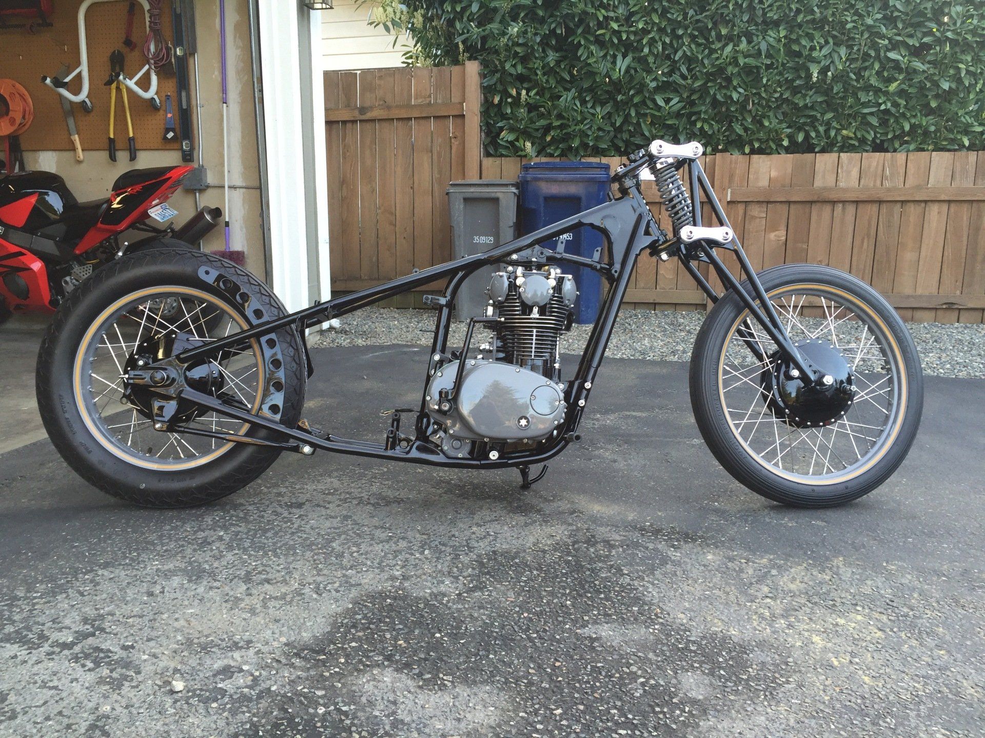 Custom blue chopper motorcycle parked on a driveway beside a garage and wooden gate