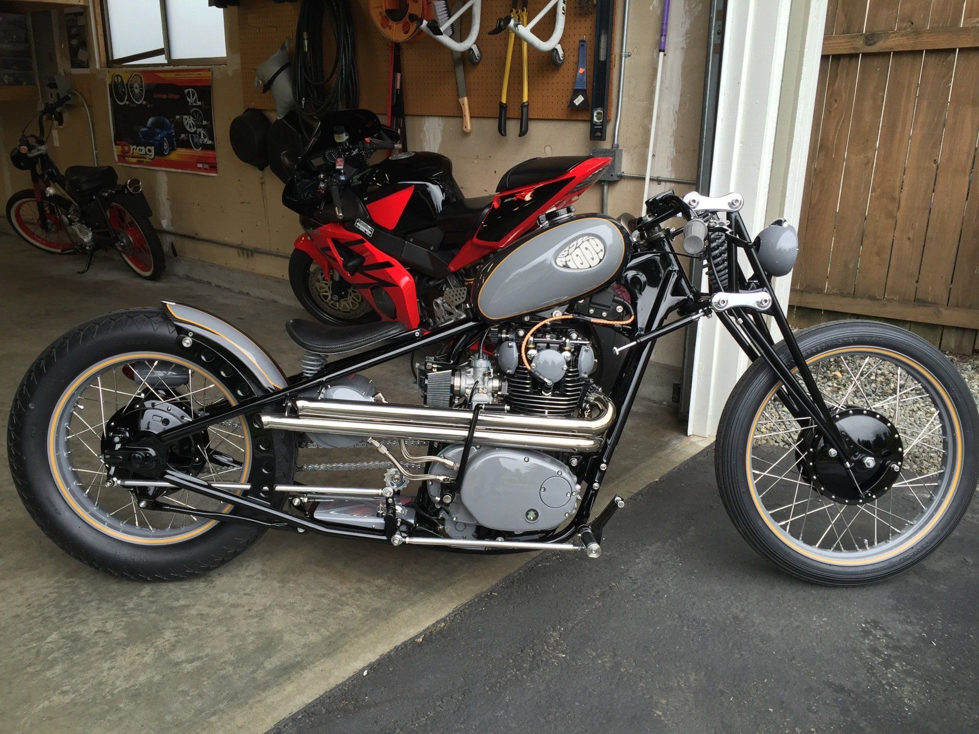 Custom silver motorcycle parked in a garage, with a red sport bike in the background.