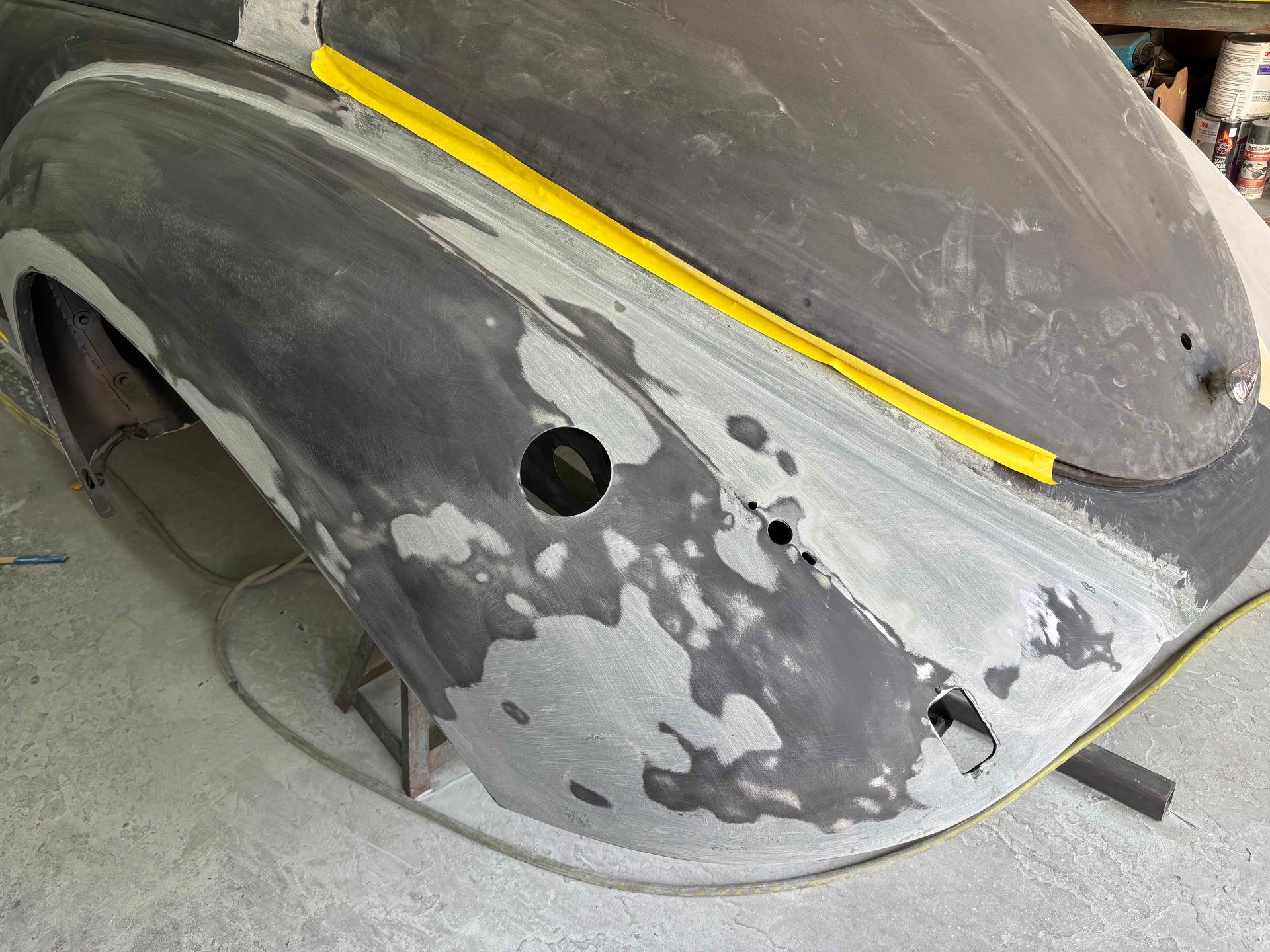 Primered car body panel with gray filler patches and a yellow masking strip in a workshop