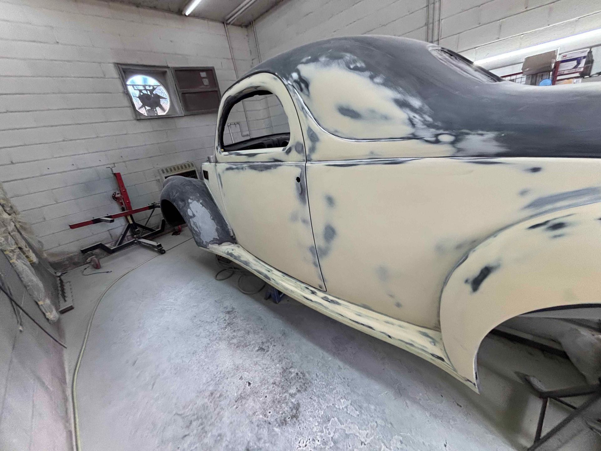 Partially restored vintage car body in a small auto body shop booth, primed cream and gray.
