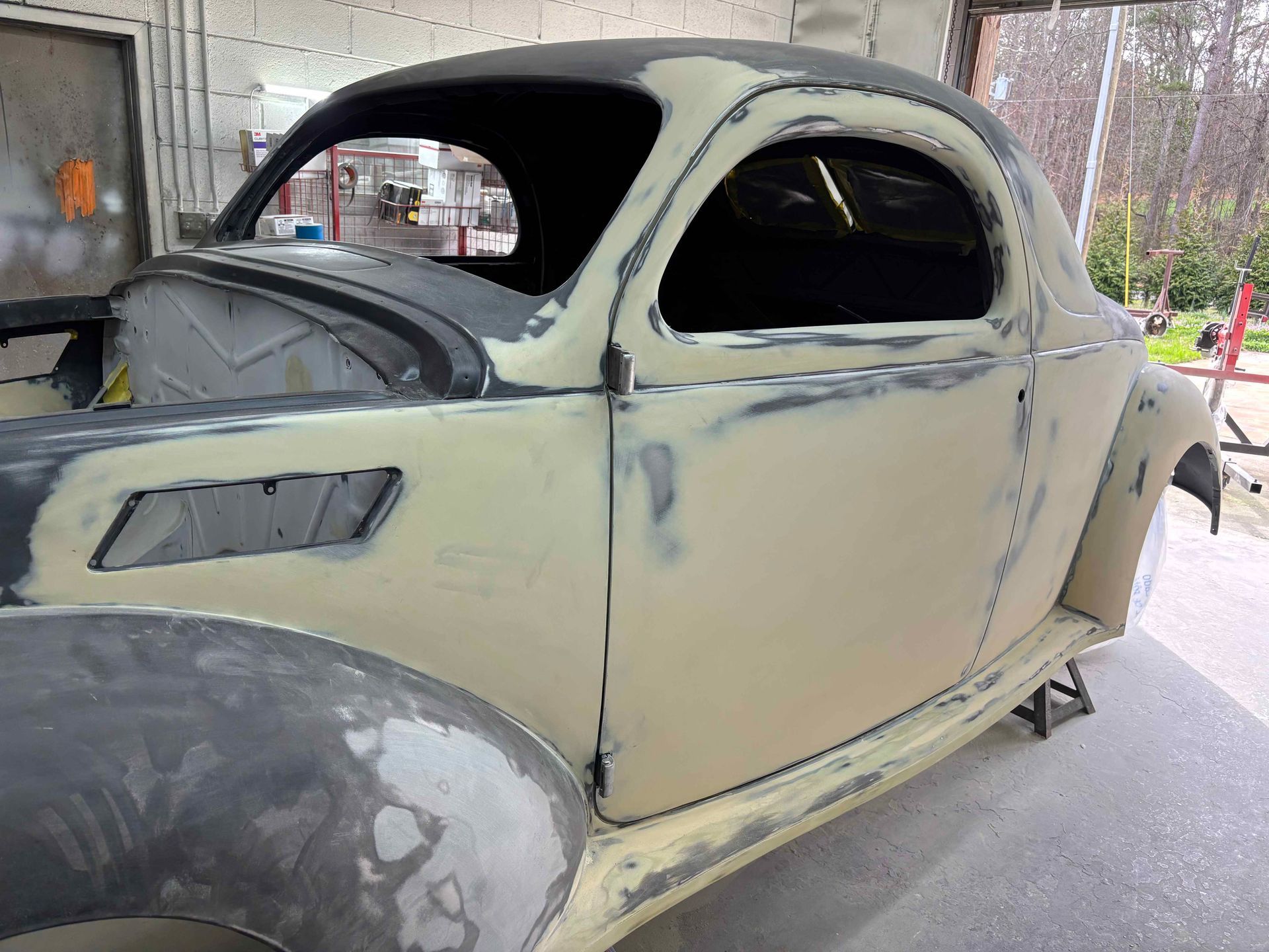 Old beige classic car body under repair in a garage, with primer-coated fenders and missing trim.