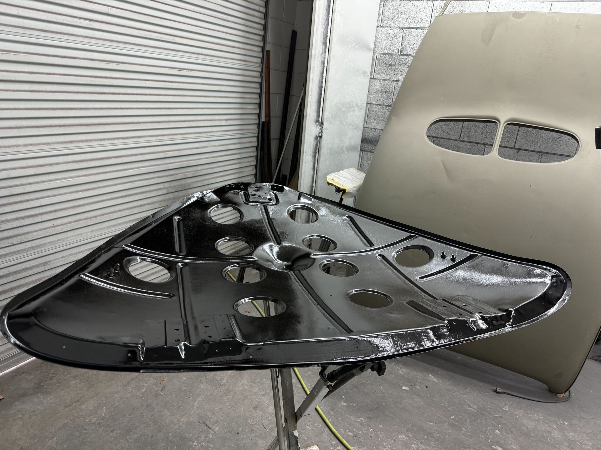 Damaged black car hood resting on a stand in a garage or workshop, with a silver panel in the background