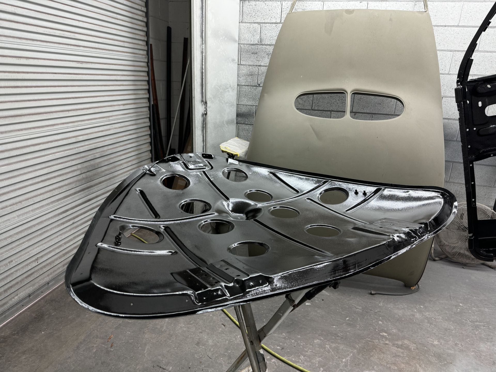 Glossy black car hood panel on a stand in a workshop, with a beige body panel behind it.