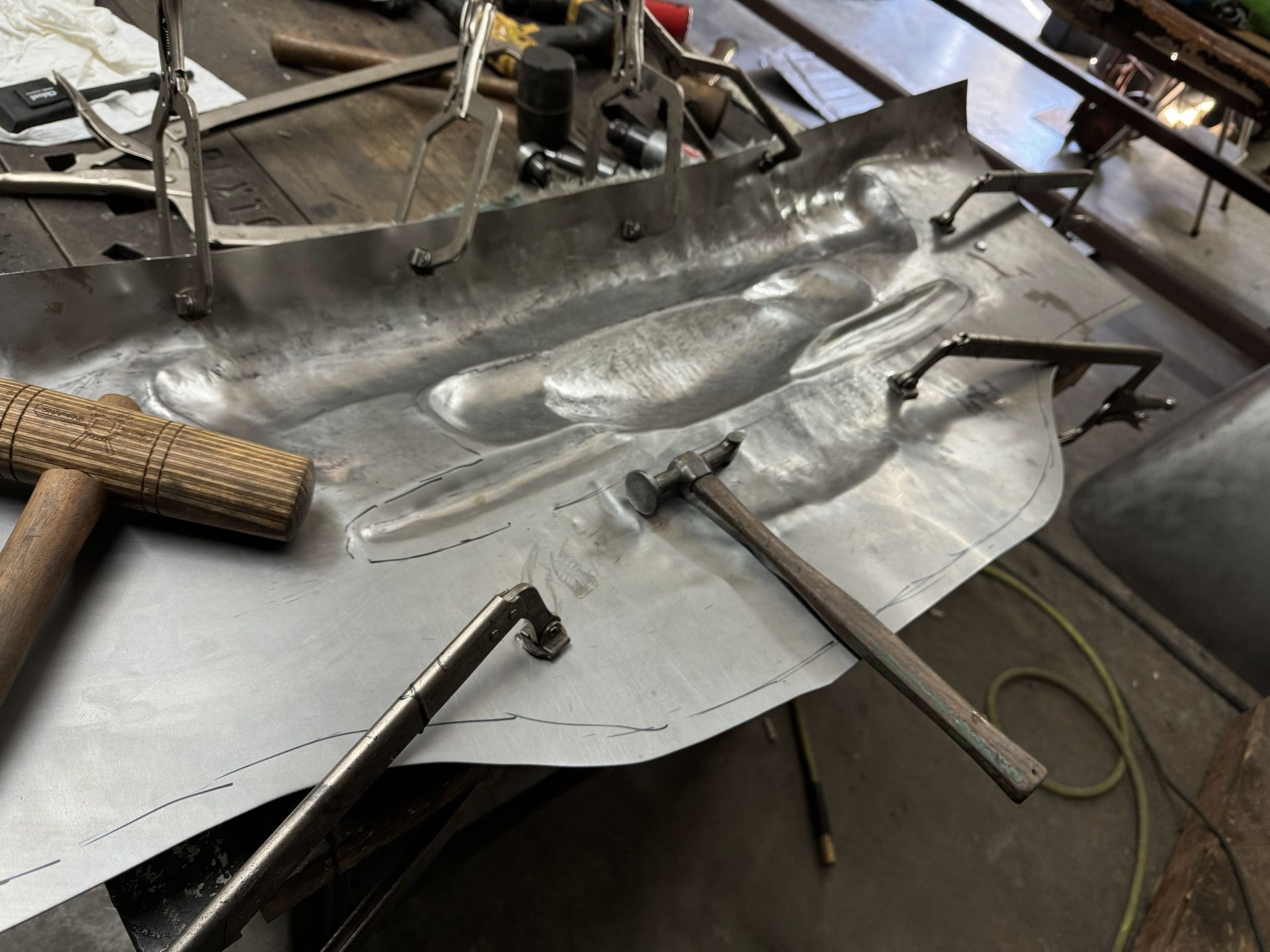 Crushed silver metal sheet in a workshop, clamped and bent on a workbench with tools around it