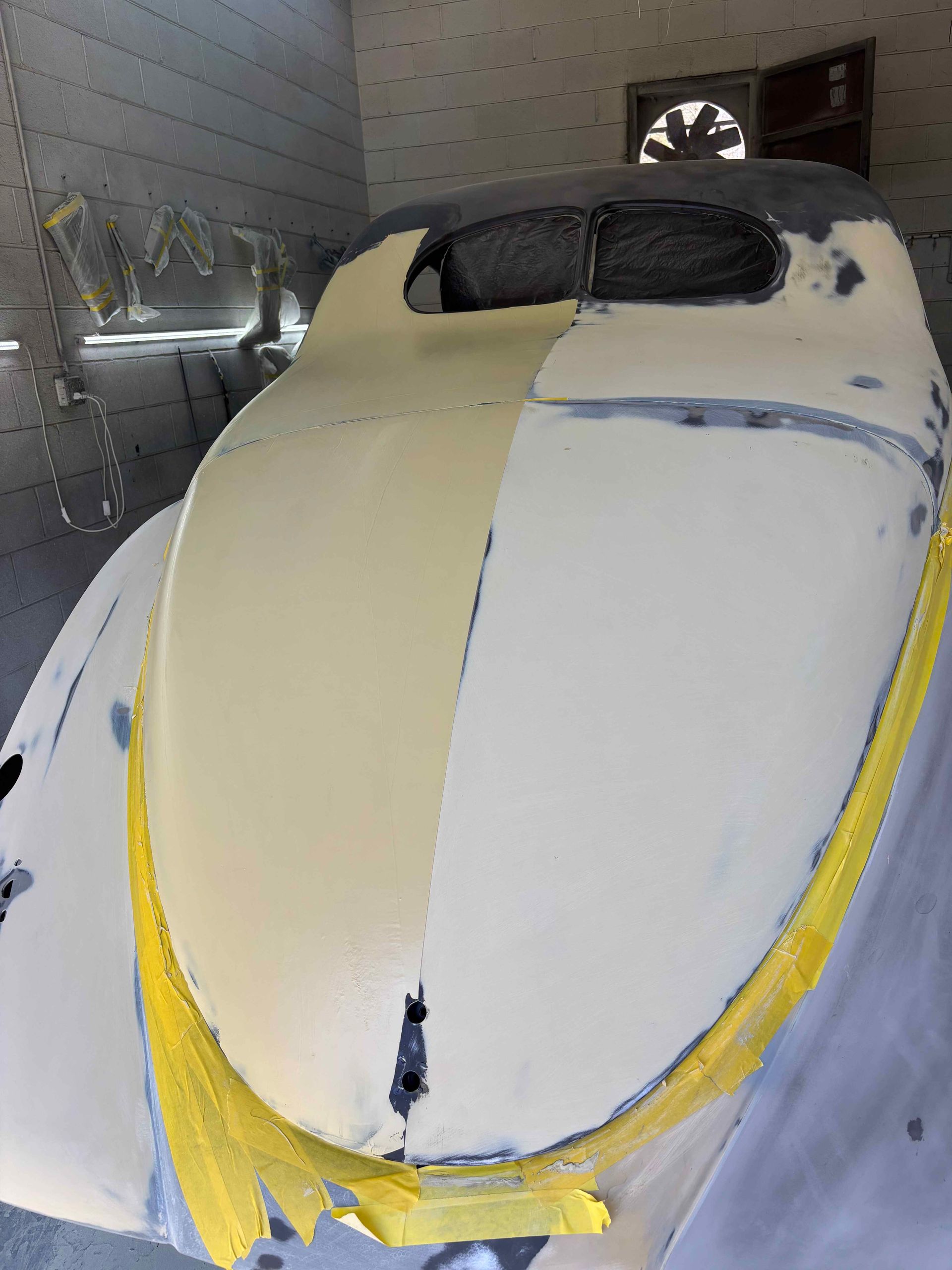 Car covered in primer and masking tape inside a garage, with a cream-colored hood panel being painted.