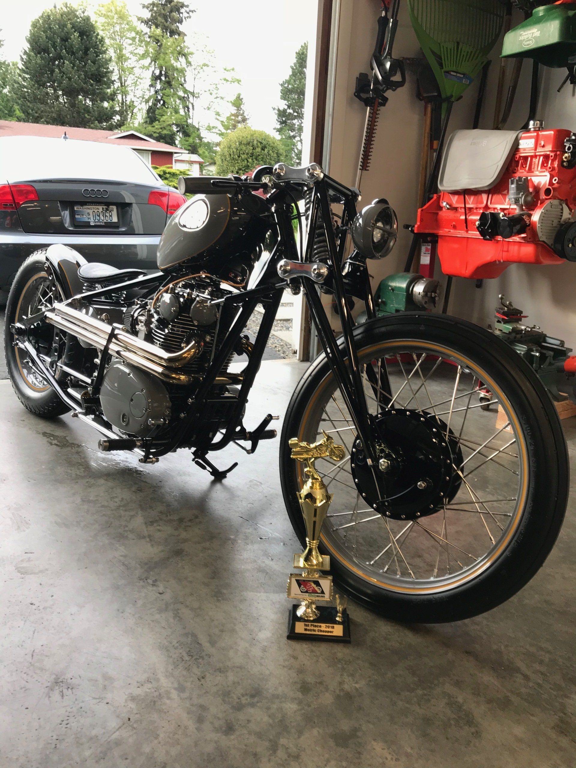 Black motorcycle with gold front wheel and trophies in a garage, parked beside a red mower and car