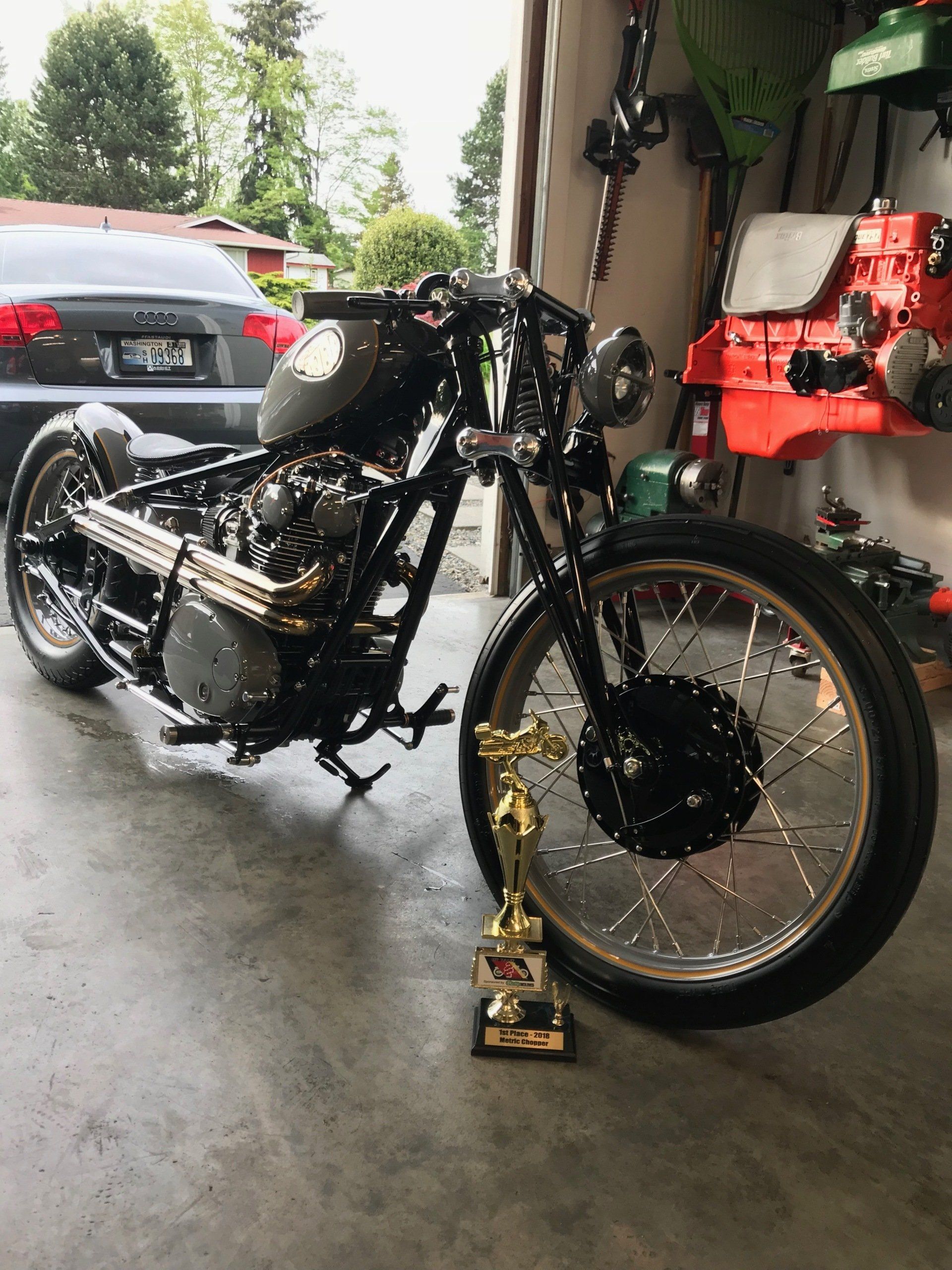 Black custom motorcycle with gold front wheel and trophy in a garage, viewed from the side.
