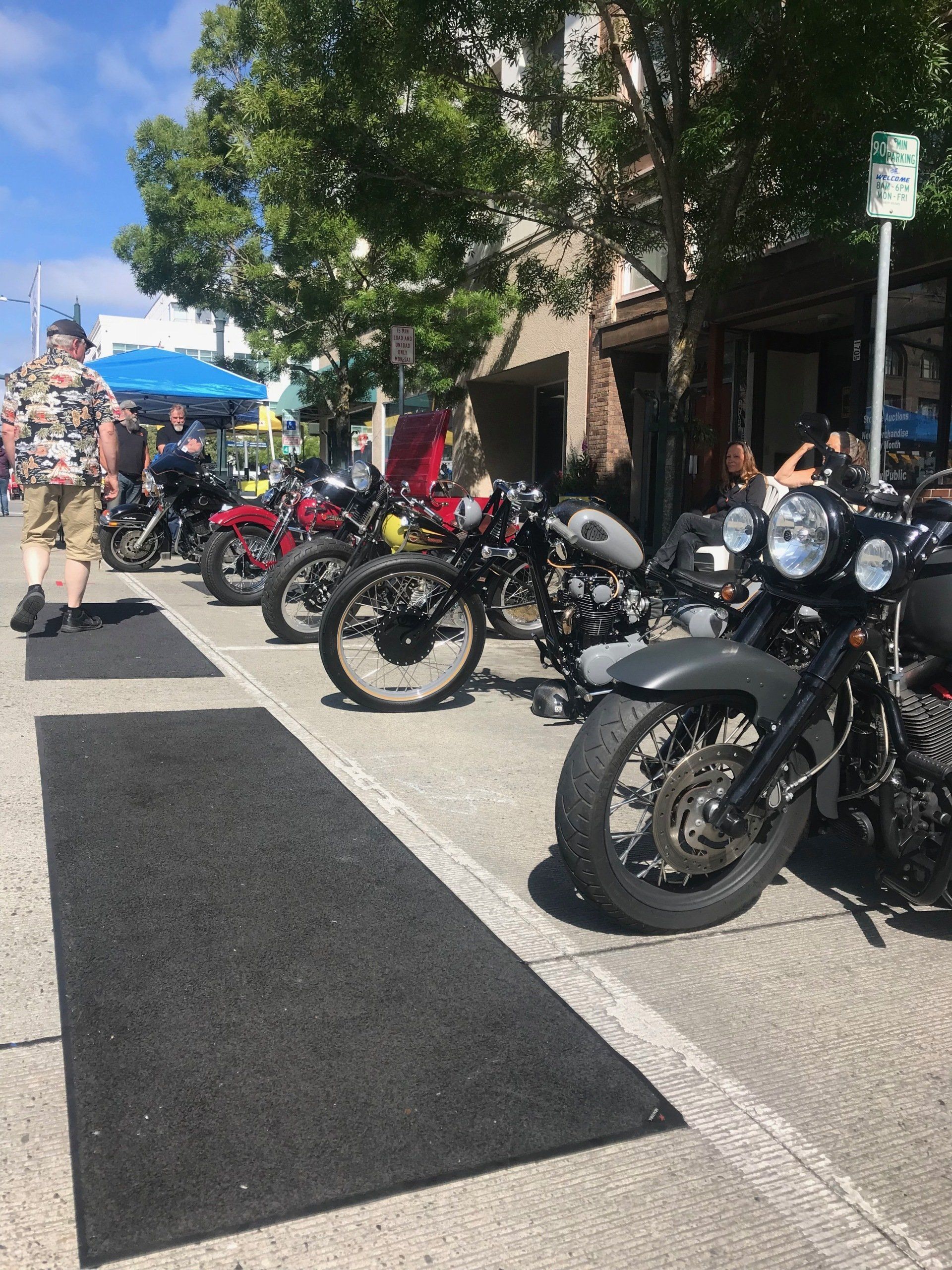 Row of parked motorcycles along a sunny sidewalk beside a building and trees
