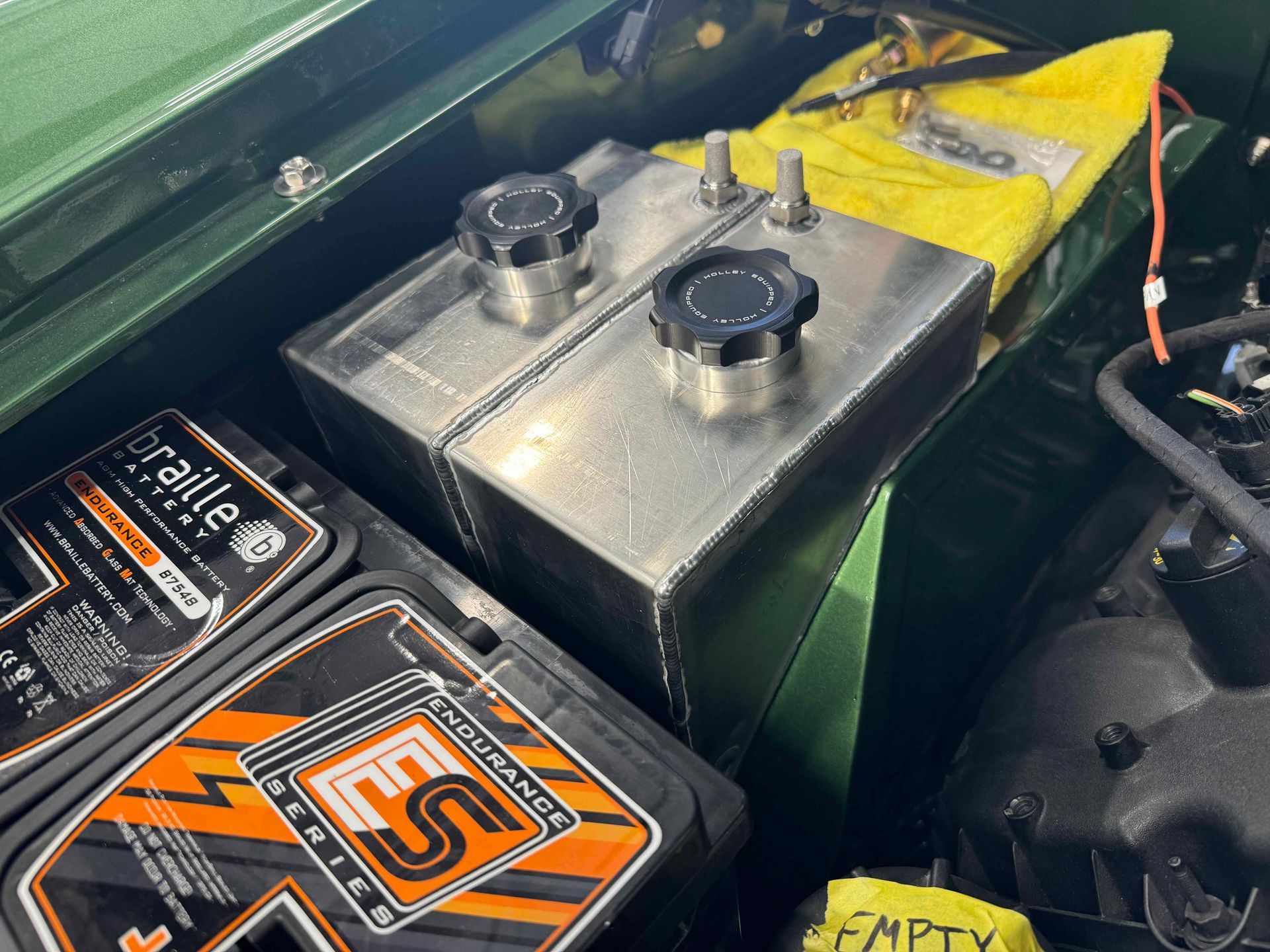 Metal fuel tank with two black caps in a car trunk beside a battery and yellow EMPTY label.