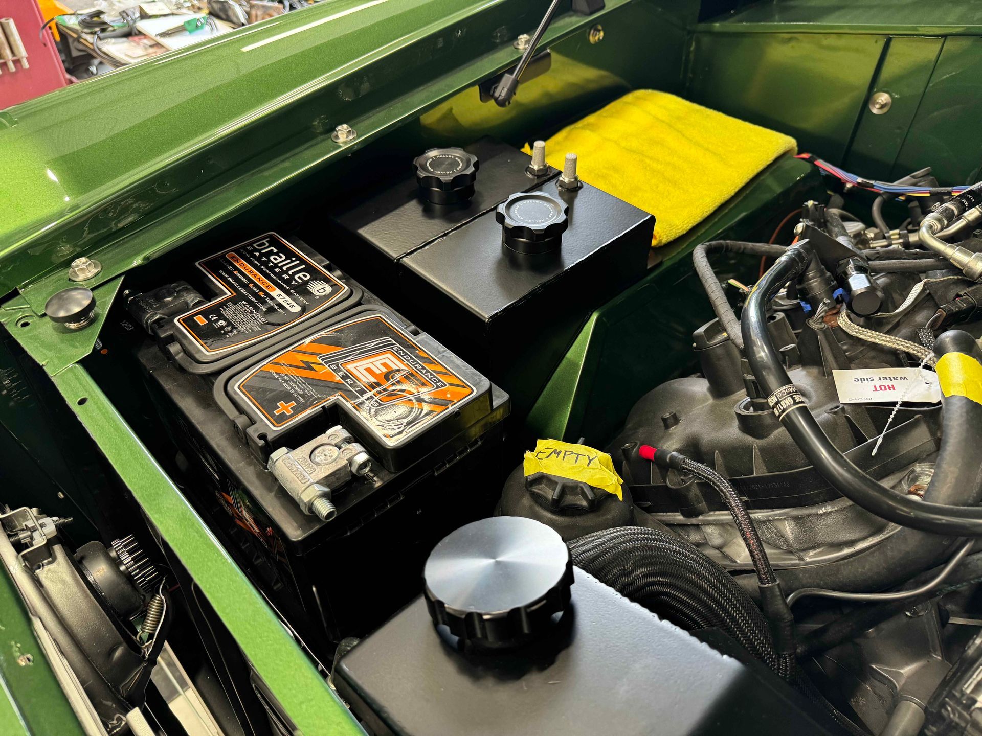 Open car engine bay with black battery, yellow cover, and hoses on a green vehicle.