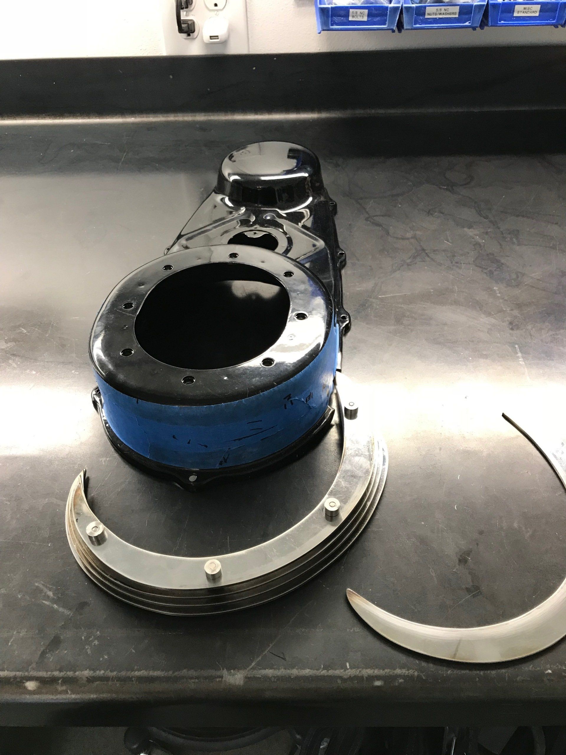 Blue and black mechanical part on a dark workbench with bolts and a curved metal piece nearby
