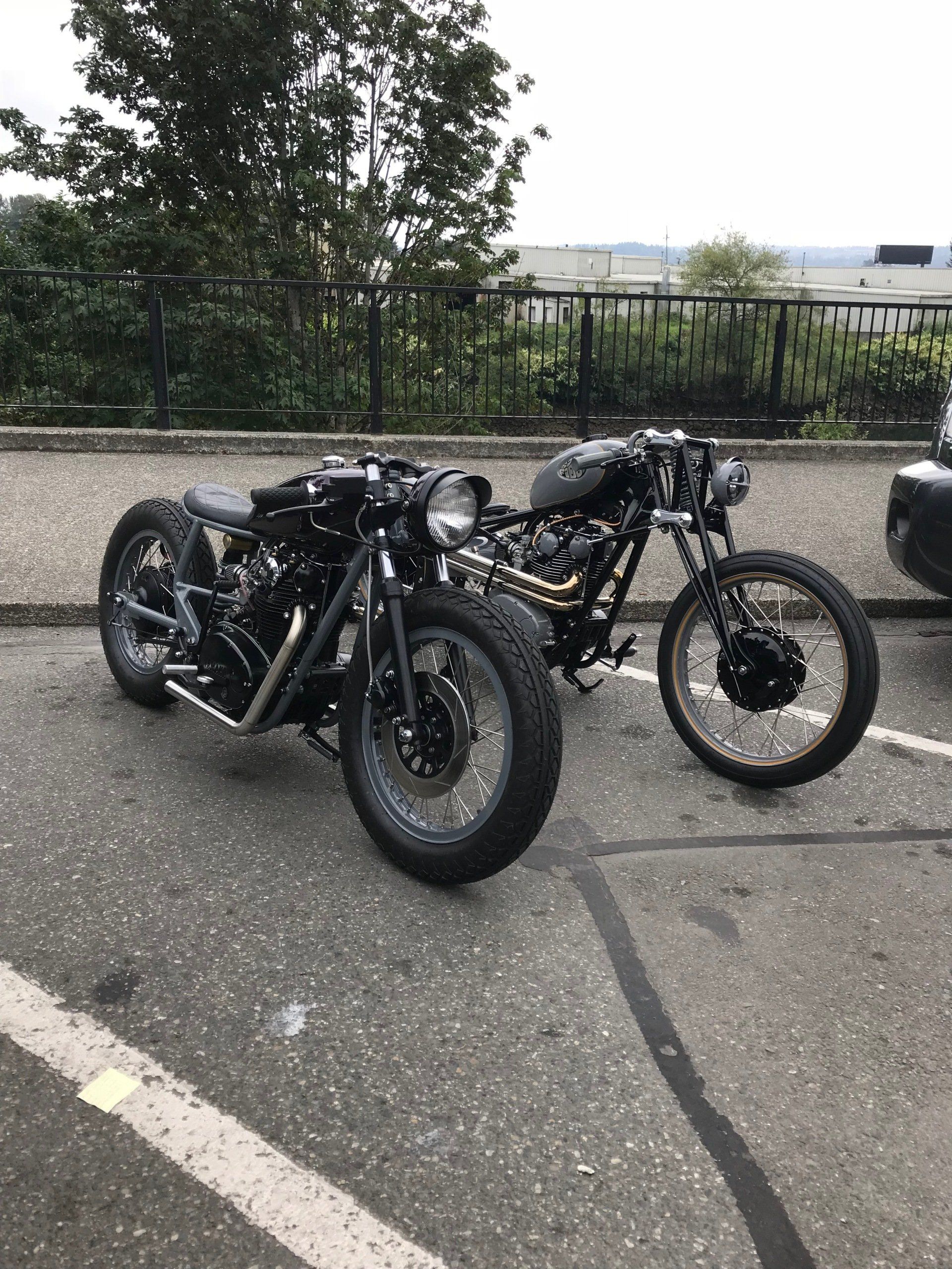 Two black custom motorcycles parked on pavement beside a fence and trees