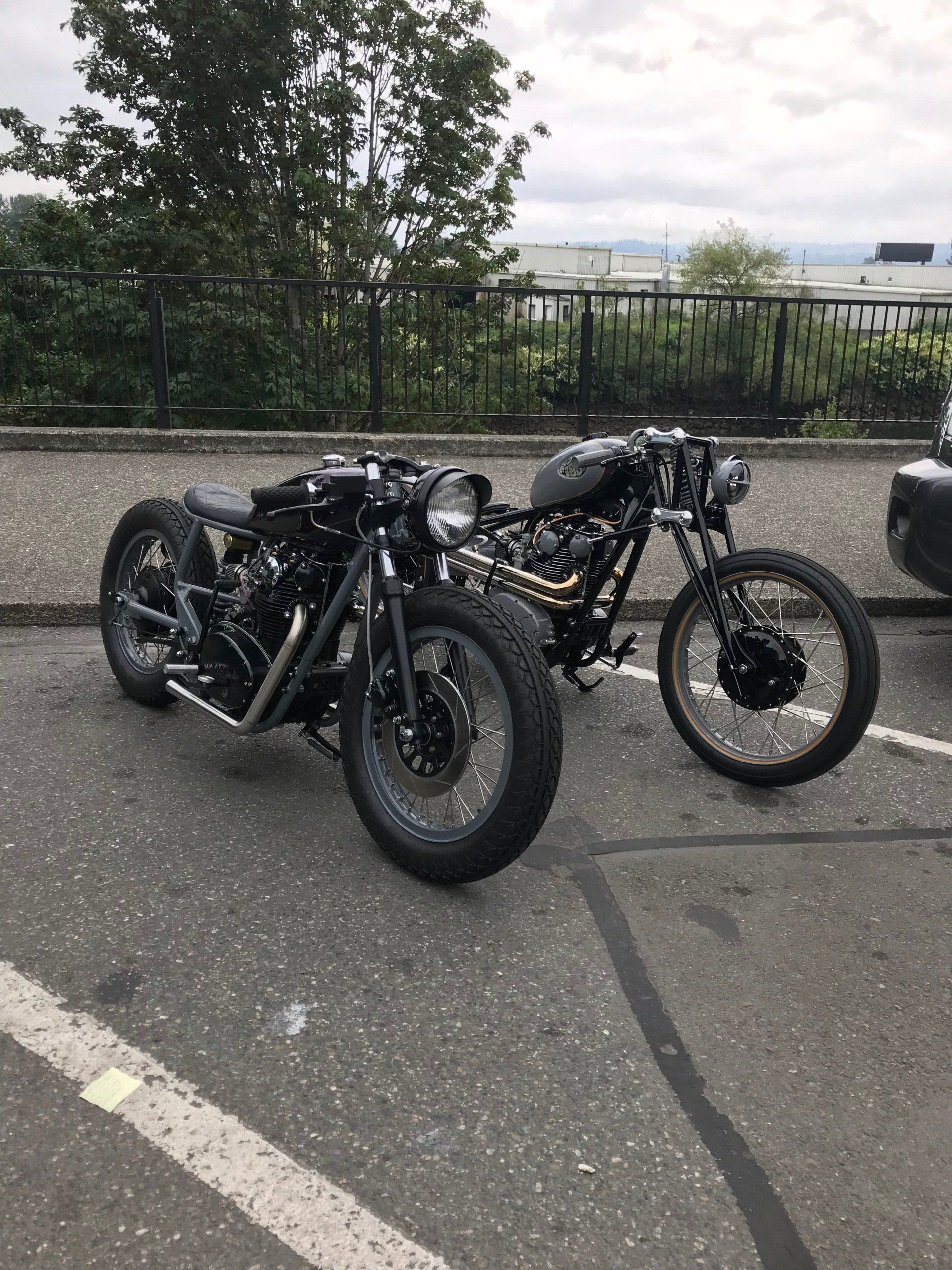 Two custom black motorcycles parked on wet pavement near a fence and trees