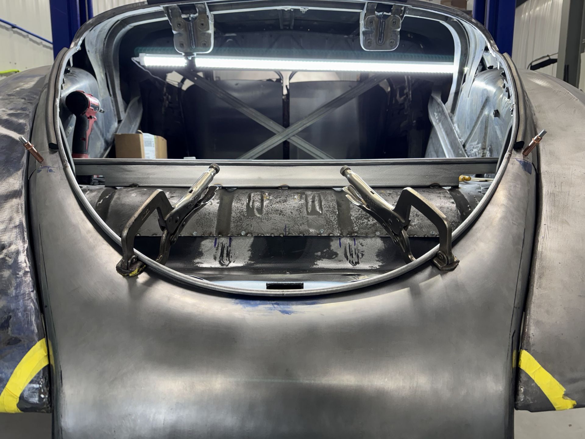 Bare car chassis in a garage, with black roll cage and unfinished gray body panels.
