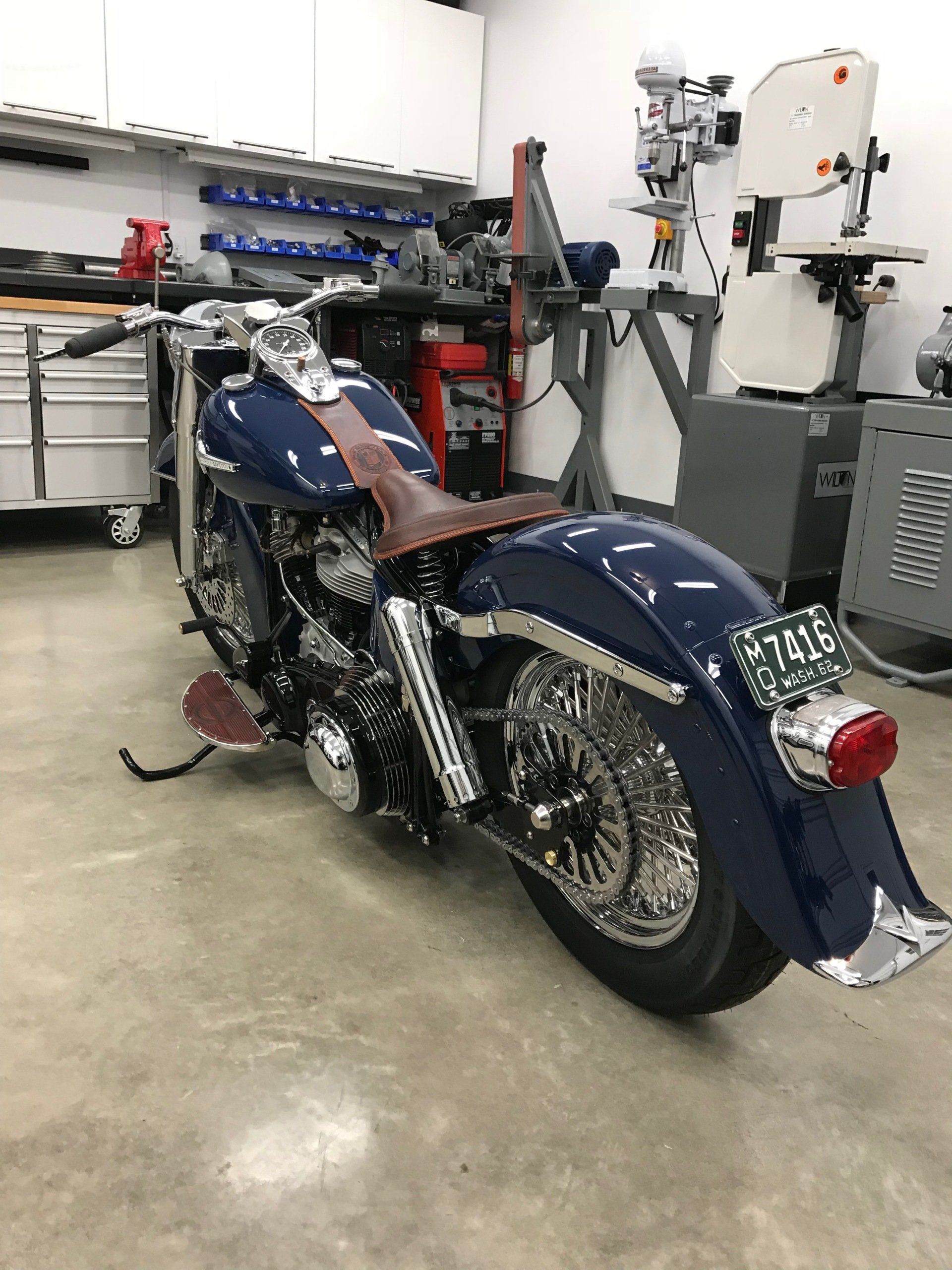 Blue motorcycle with brown seat in a workshop, shown from the rear side.