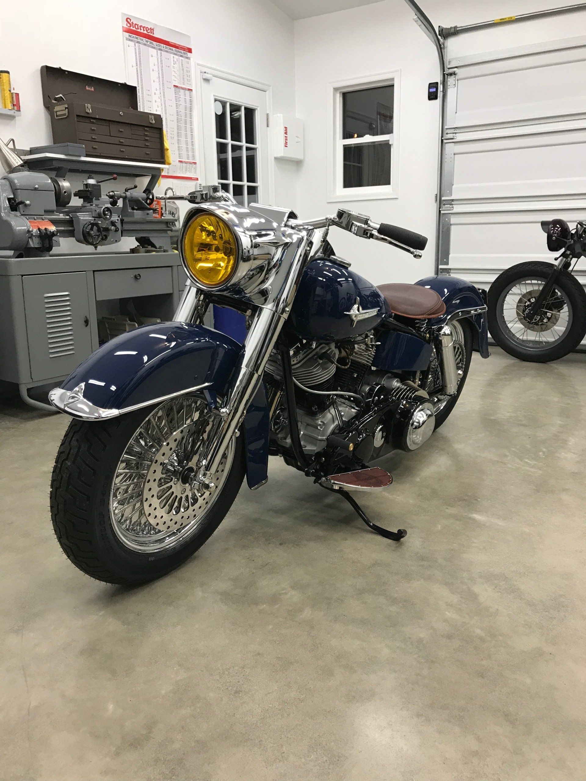 Blue motorcycle with a yellow headlight in a garage, with another bike and tools in the background