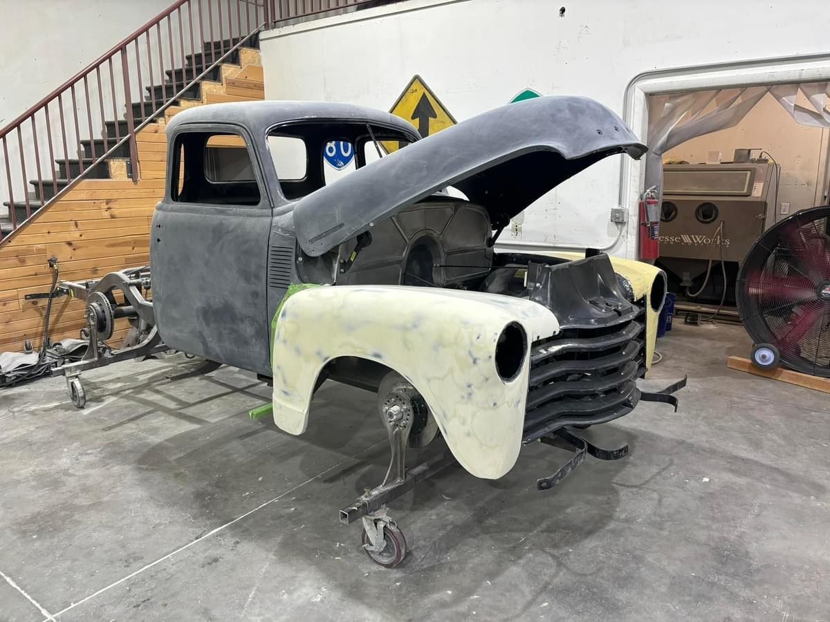 Partially restored vintage pickup truck in a garage, hood open and front fender removed.