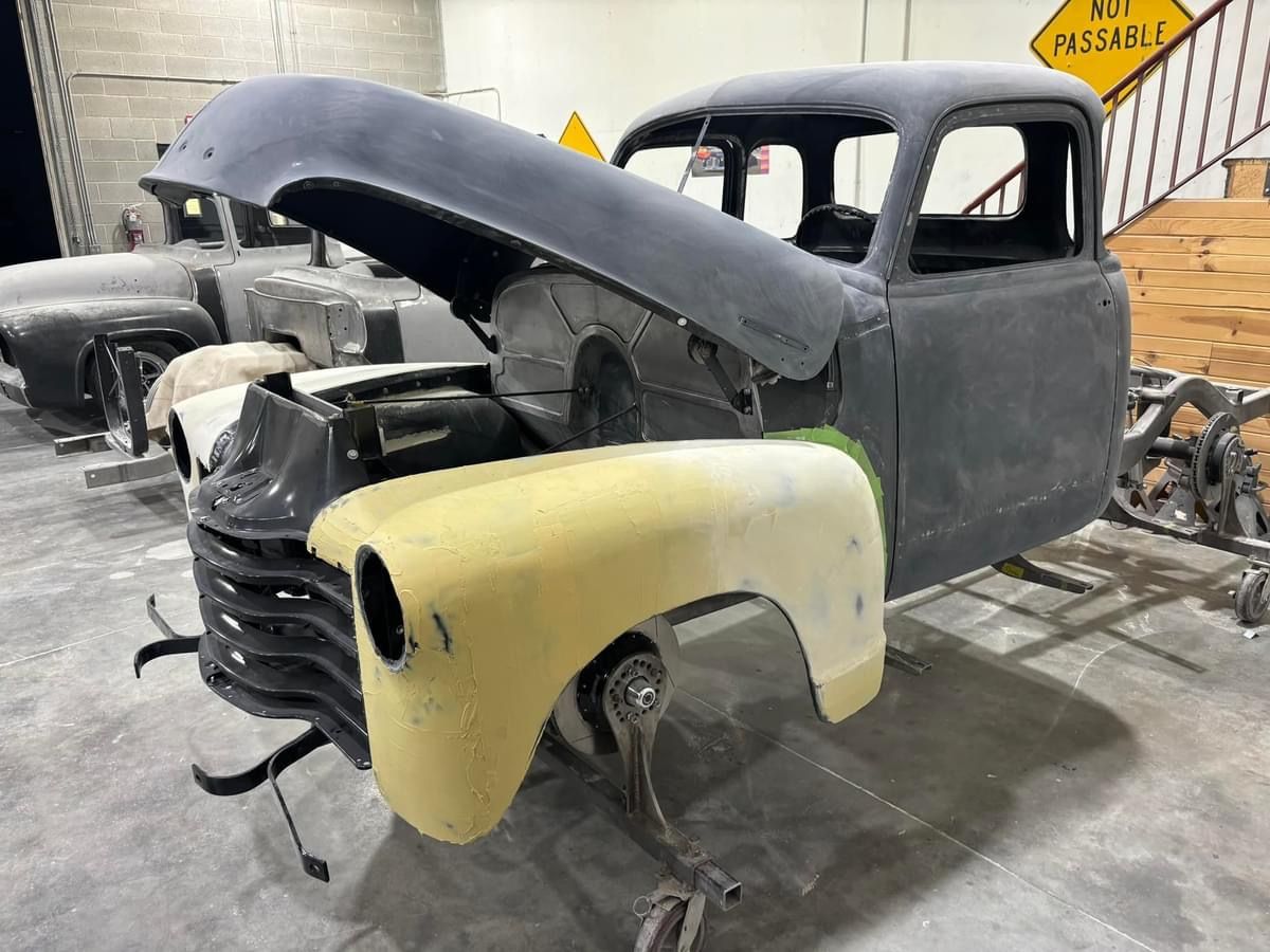 Rusty vintage truck body in a workshop, with a yellow front fender and raised hood
