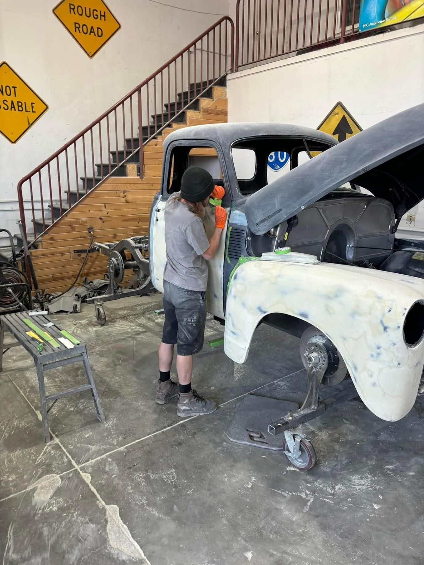 Person working on a white car in a garage, with a black hood open and a staircase in the background