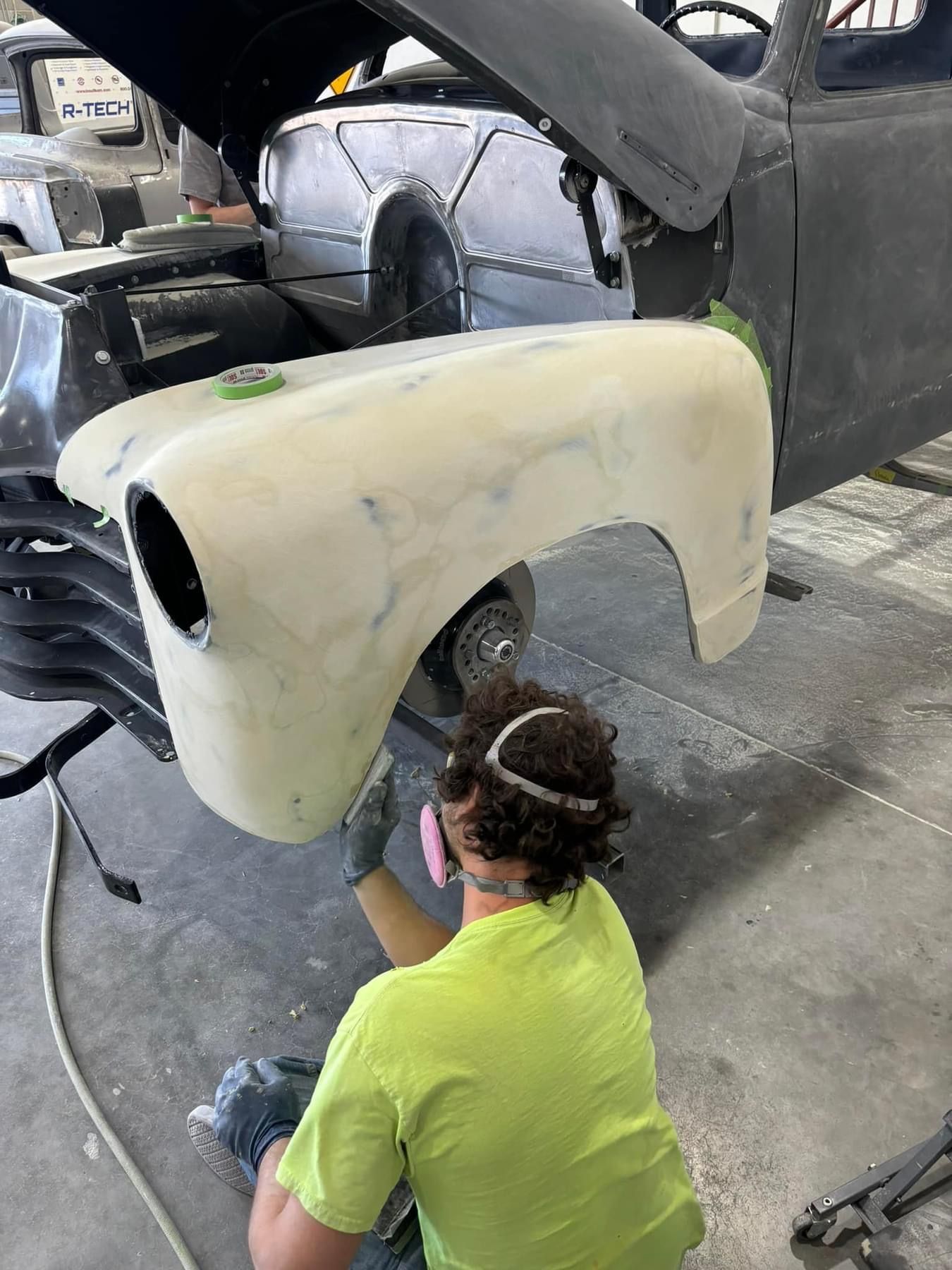 Person in neon shirt sanding a white car body panel in an auto repair shop