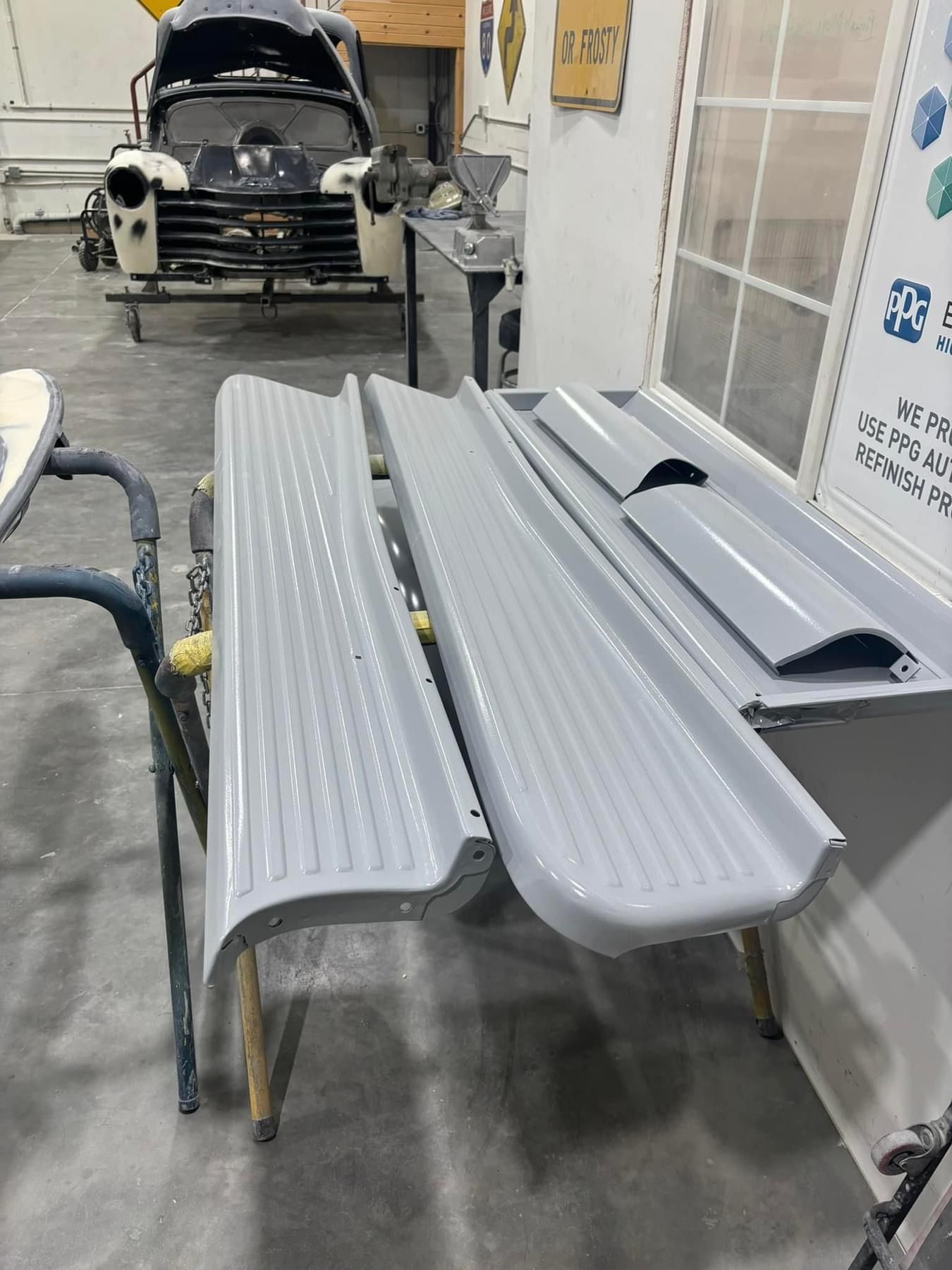 Grey corrugated truck bed panels on a workbench in an auto body shop, with a black truck in the background
