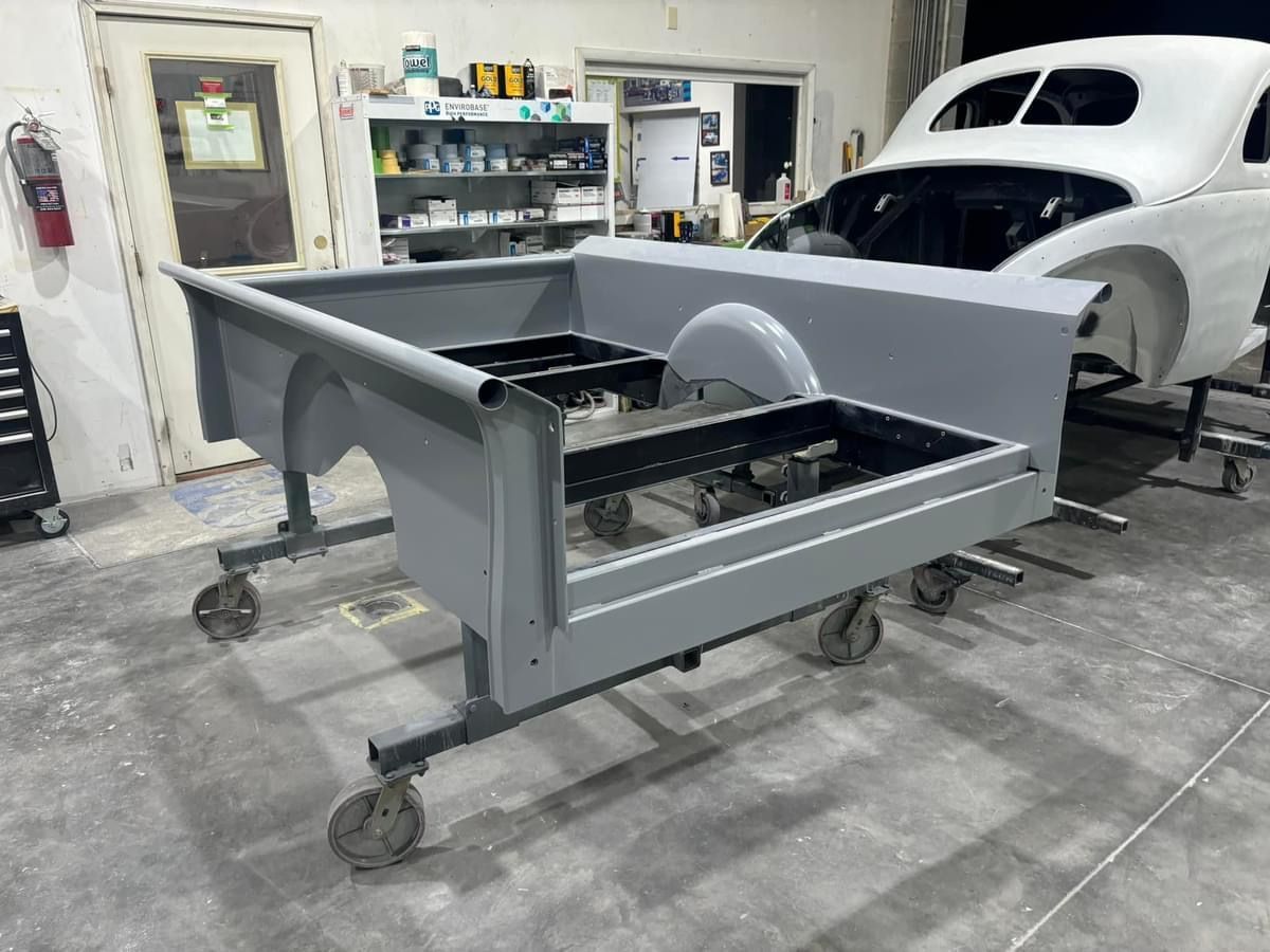 Gray metal car chassis on casters in a workshop, with a white vintage car body in the background.