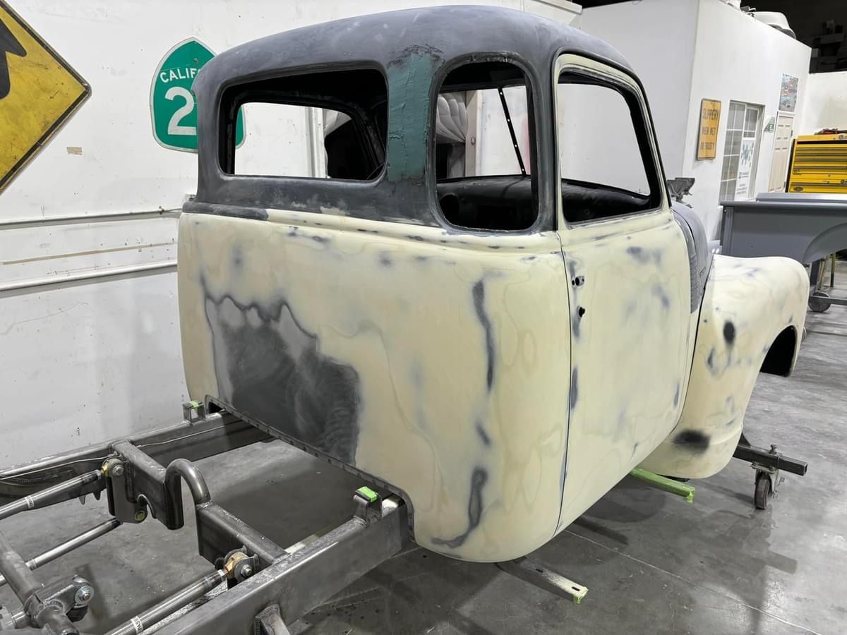 Primered vintage truck cab on a workshop frame, showing sanded bodywork and blacked-out windows.
