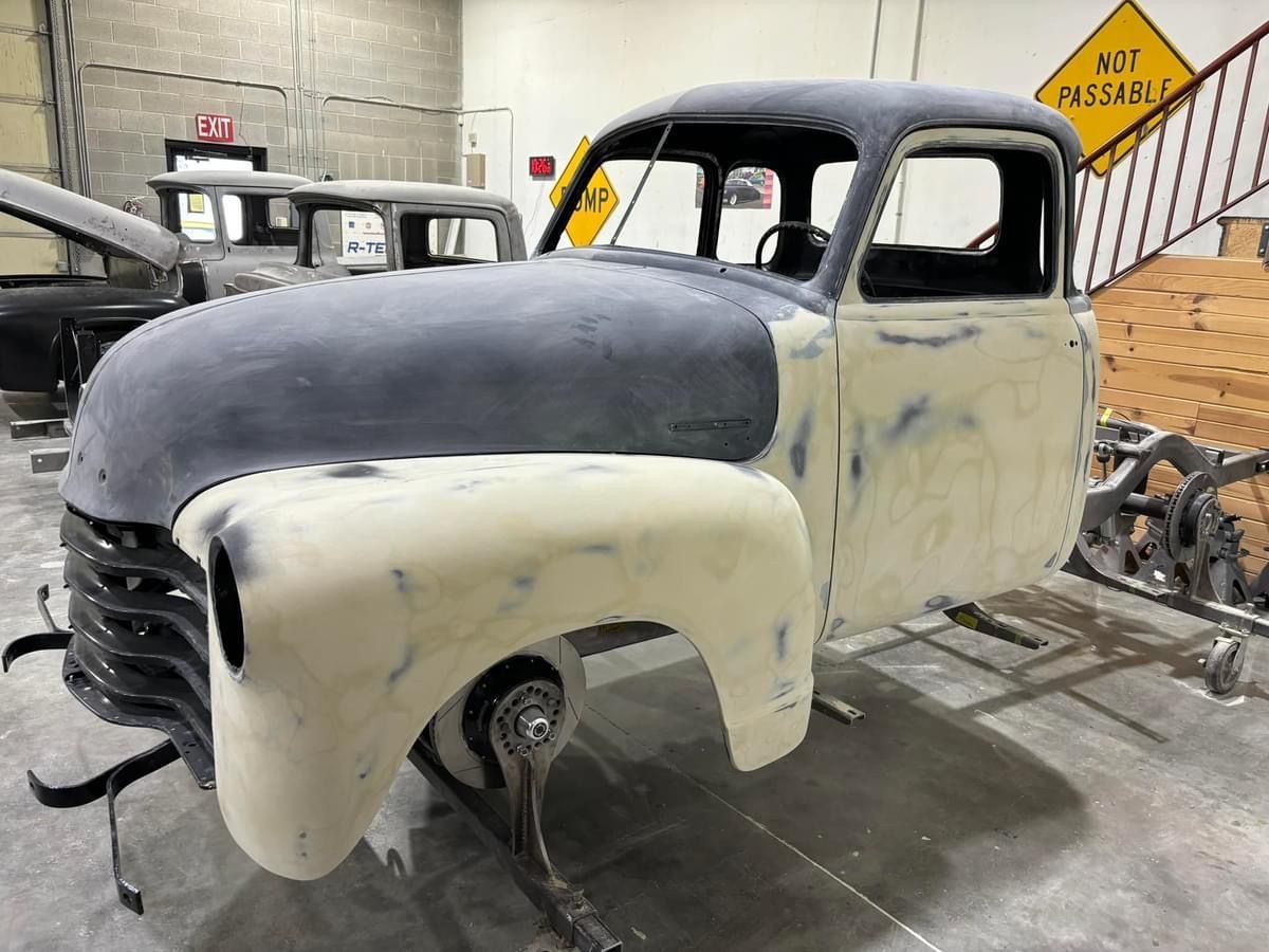 Primer-painted vintage pickup truck body in a workshop, black hood and cream fenders, missing wheels.