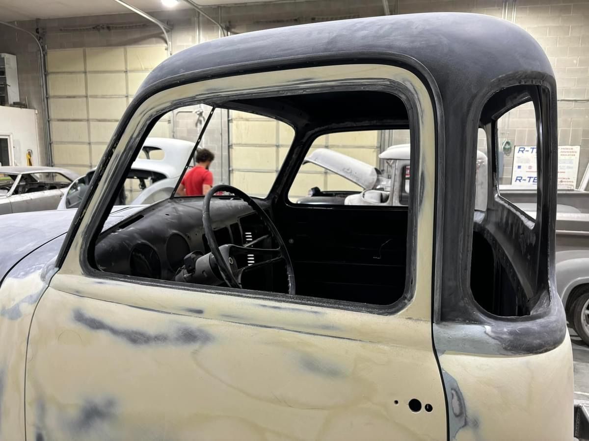 Primer-gray vintage truck cab in a workshop, with windows and doors missing.