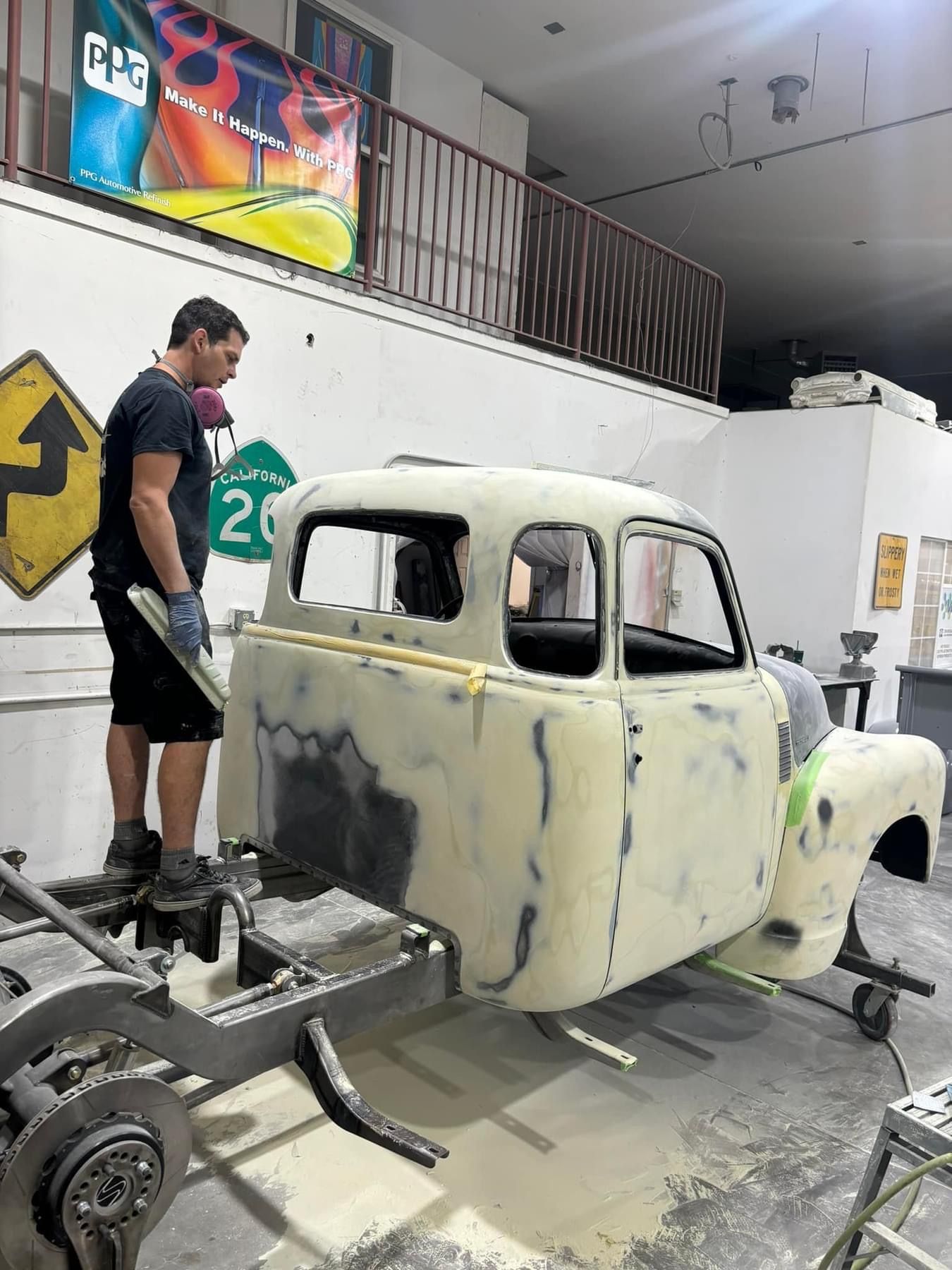 Man working on a vintage car body in a garage, with a bare chassis and primer patches.